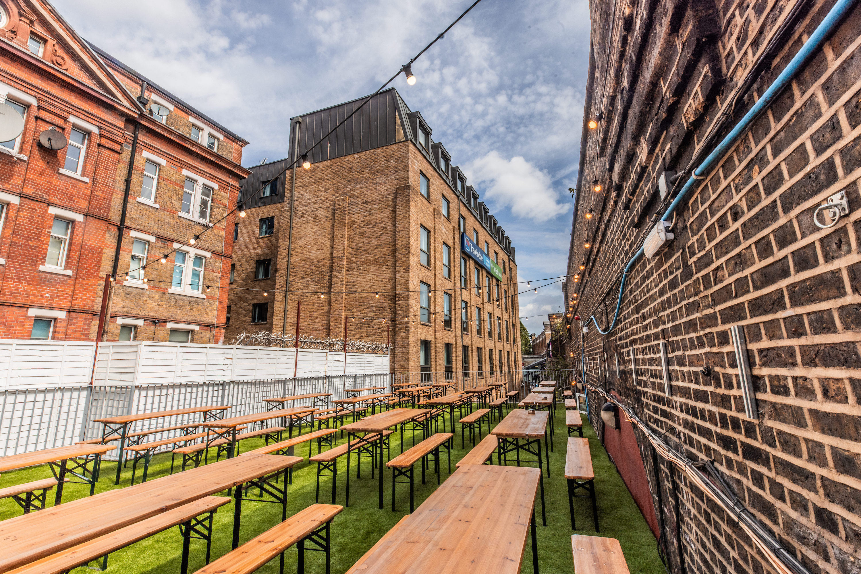 A view of the Vauxhall Food and Beer Garden for 800, showing rows of picnic tables on green turf, an exposed brick wall, and string lights.