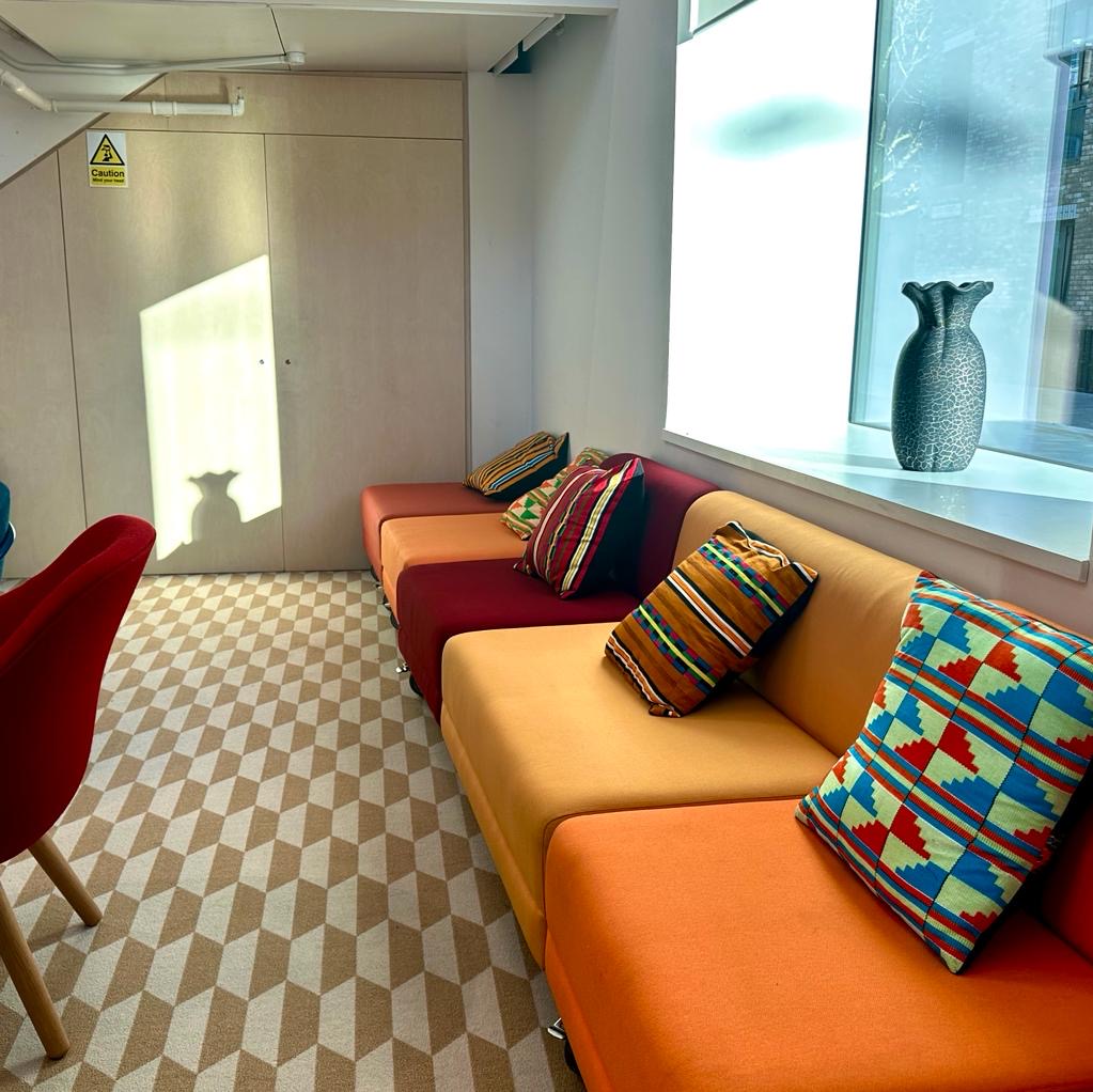 A sunlit corner of the 40-capacity Lounge at Melanin Health & Wellness, featuring a colorful orange and red sofa with vibrant, patterned throw pillows