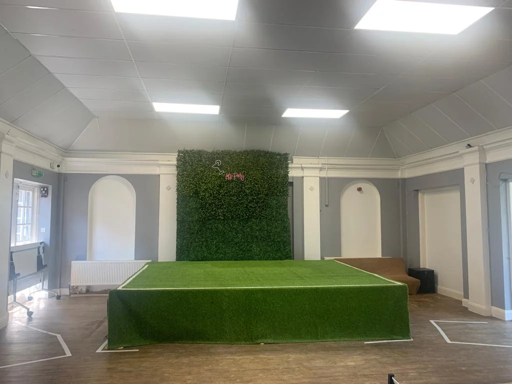 A view of the function room with a large, elevated stage covered in artificial turf, in front of a tall, green hedge wall with a neon sign.
