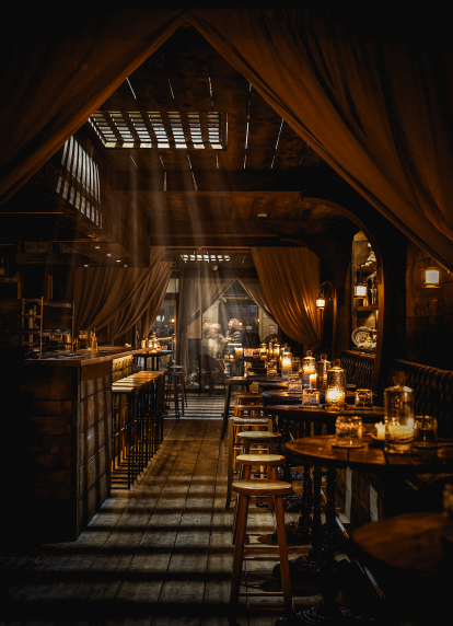 Interior view of a rustic, candlelit tavern featuring a long wooden aisle, high tables with stools, and dramatic shafts of light beaming through ceili