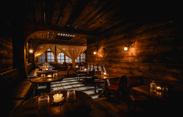 An intimate seating corner in a wooden tavern with dark paneling, warm wall-mounted lanterns, leather bench seating, and small wooden tables illuminat
