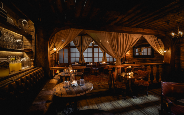 A cozy, vintage-style seating area separated by a wooden railing, featuring shelves lined with glass jars, cream-colored draped curtains, and a rustic
