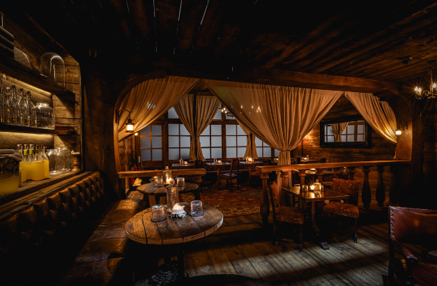 Atmospheric view of a tavern interior showing a leather banquette, rustic wooden tables, and a raised seating area framed by theatrical drapes and dar