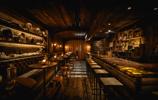 A long, dark wooden bar featuring a unique illuminated amber countertop, lined with metal stools, situated opposite a leather bench seat with shelves 