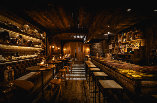 Wide shot of the main bar area in The Rhum Tavern, highlighting the rustic wooden floor, a fully stocked back bar, and warm ambient lighting reflectin