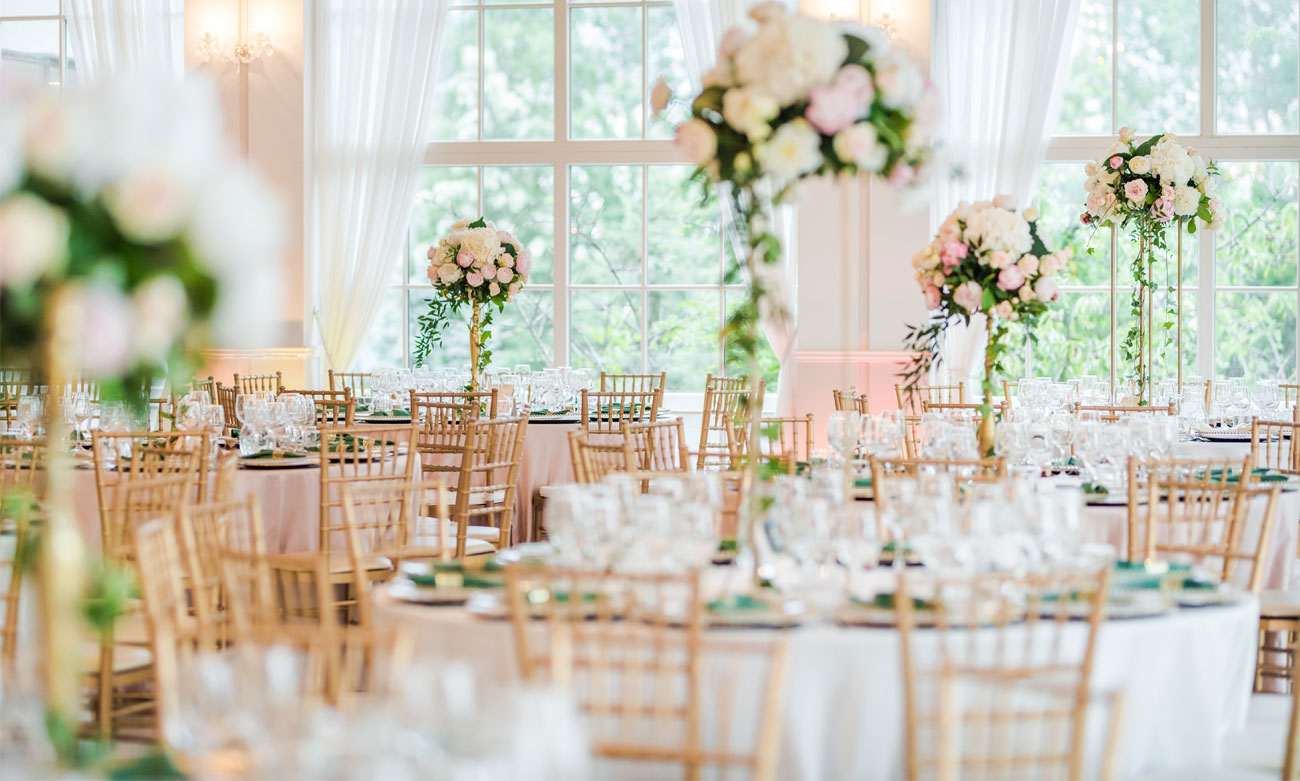 Elegant wedding reception at The Gulbenkian featuring round tables, floral centerpieces, gold chairs, and floor-to-ceiling garden views.