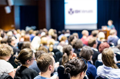 Large audience seated for a conference at ISH Venues, with a speaker at the podium in front of a projection screen.