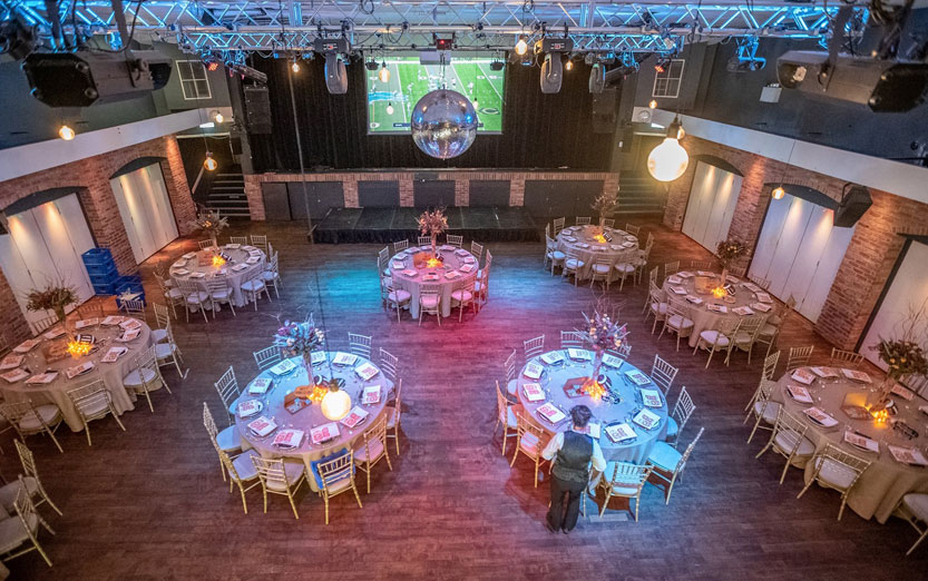High-angle view of Venue 1 at ISH Venues showing a banquet layout, large disco ball, professional lighting rig, and a stage with a sports broadcast.