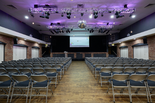 Wide shot of Venue 1 in theater layout with rows of chairs facing a stage, screen, and professional lighting rig.