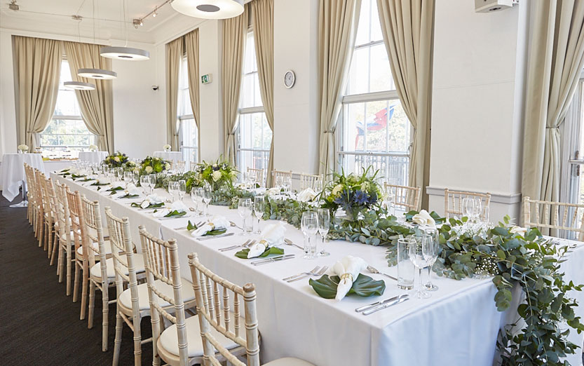 Elegant banquet dining setup with long floral-decorated tables, cream curtains, and natural light from large Marylebone park-view windows.