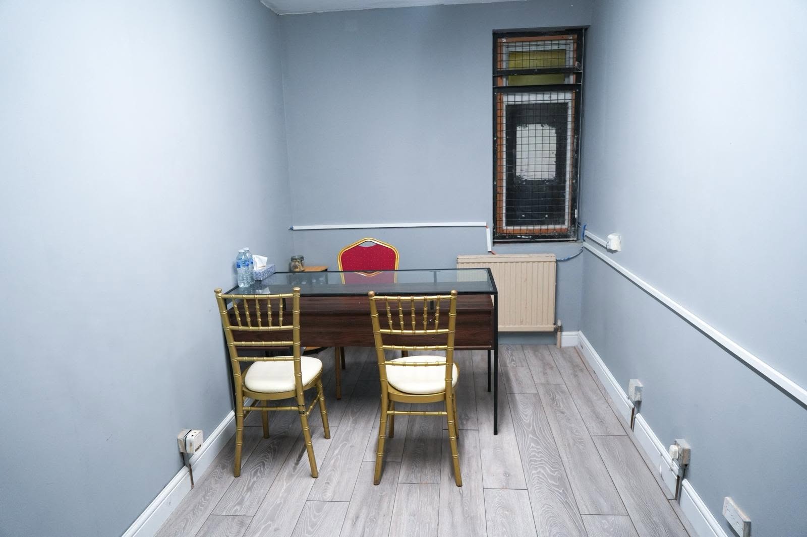 Stylish Canning Town meeting room featuring a glass-topped desk, elegant gold Chiavari chairs, and soft blue walls.