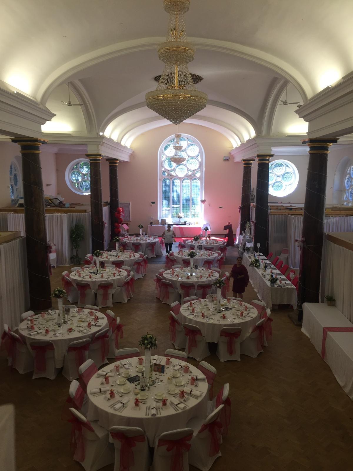 Round tables dressed with white linen and floral centrepieces fill St Matthias Church Hall, set up for a formal dinner or wedding reception beneath th