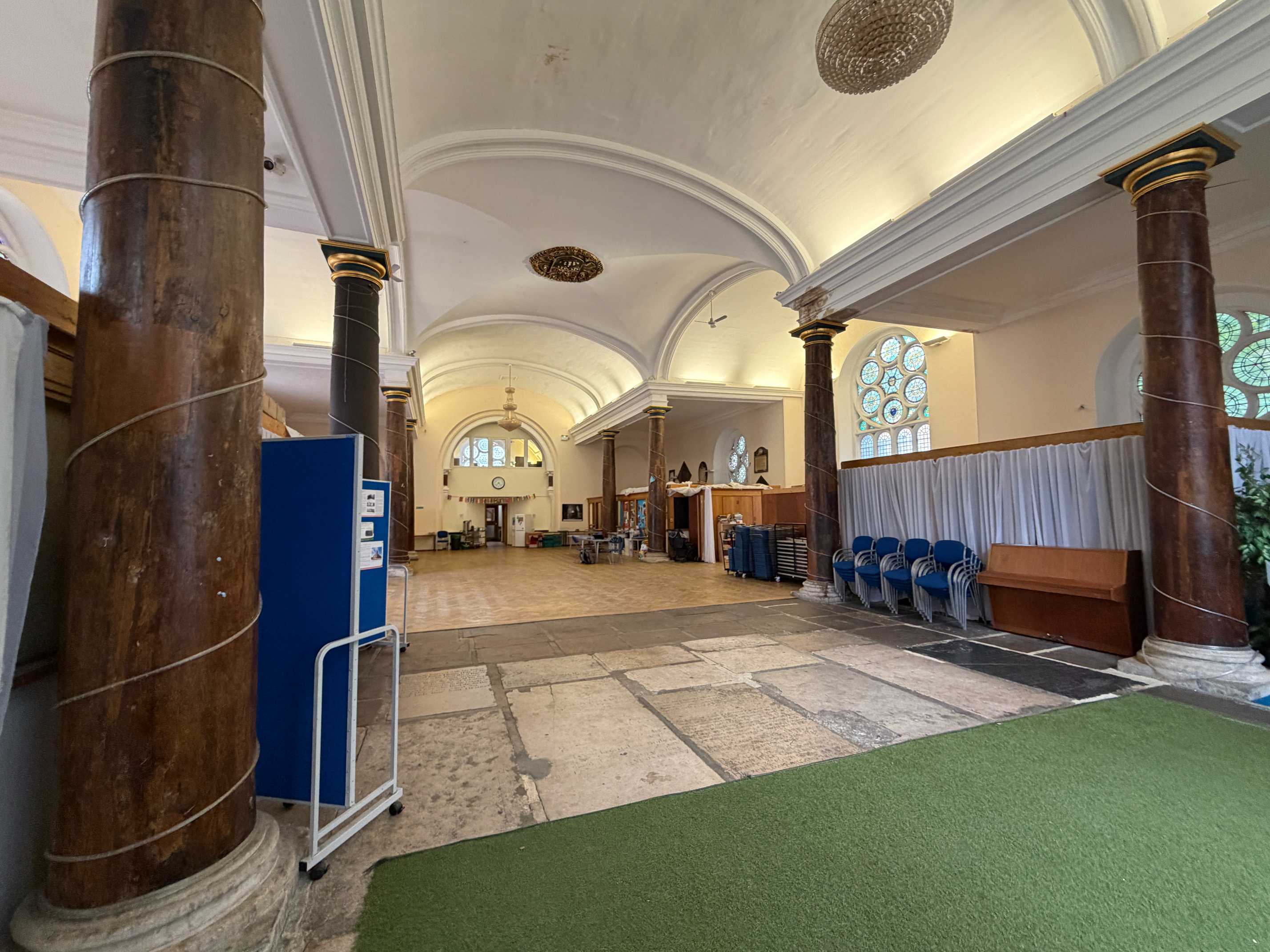 Rows of blue chairs are arranged along the side aisle of St Matthias Church Hall, framed by stone arches and stained glass windows, ready for a ceremo