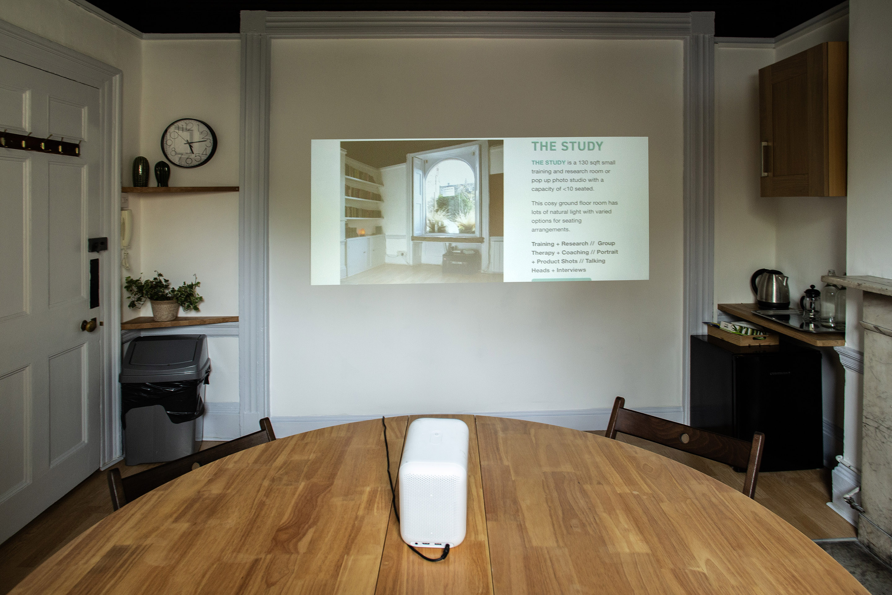 A small photography studio for 10, featuring a round table, projector screen, and a small kitchenette.