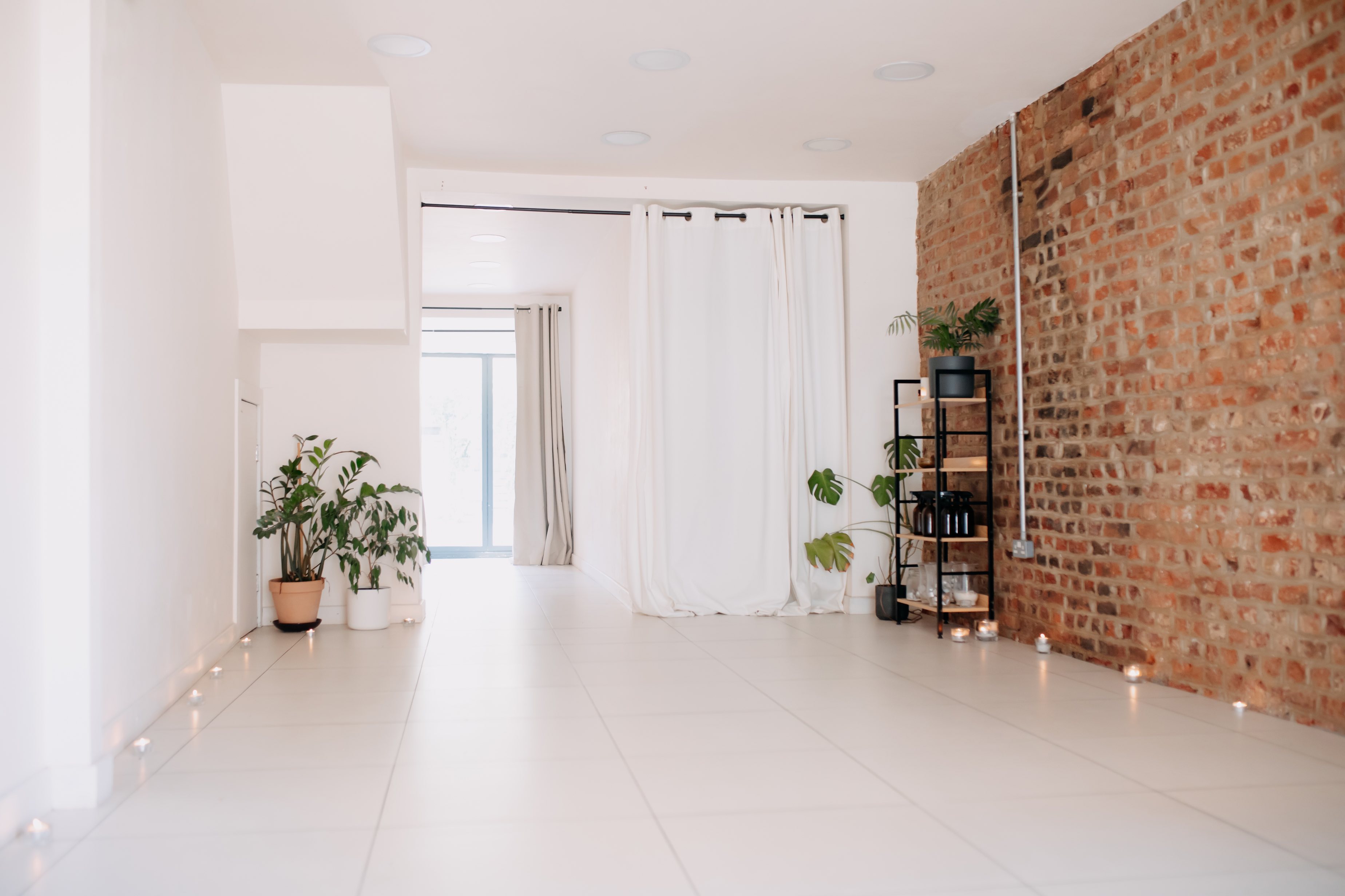 A peaceful wellness studio with a white tile floor and an exposed brick wall, featuring a shelving unit with plants and candles - book via Conveenie.