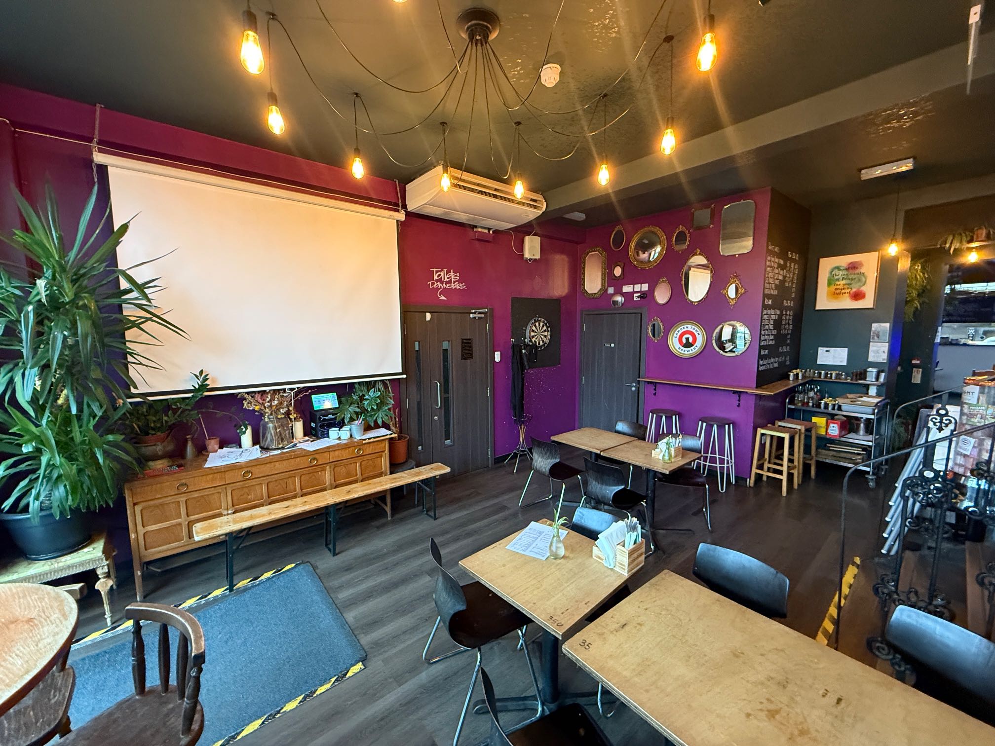 A private function room with a projector screen and a dartboard, set against a purple wall with various framed mirrors.