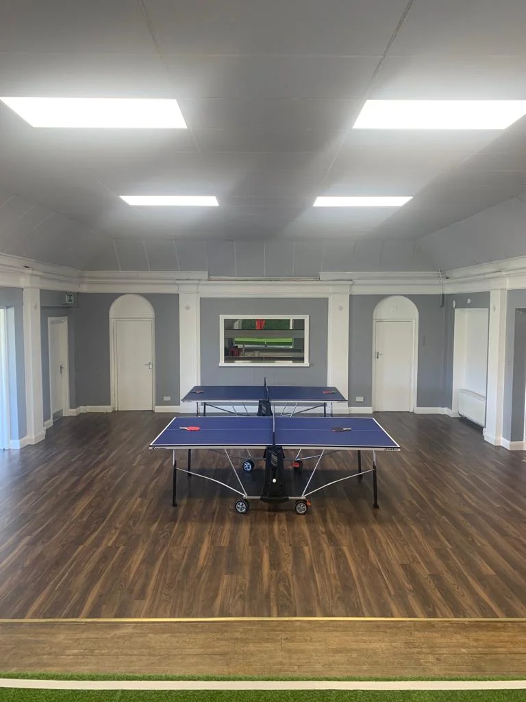 Function room with two blue ping-pong tables on a glossy wood floor, with white walls and arched doors.