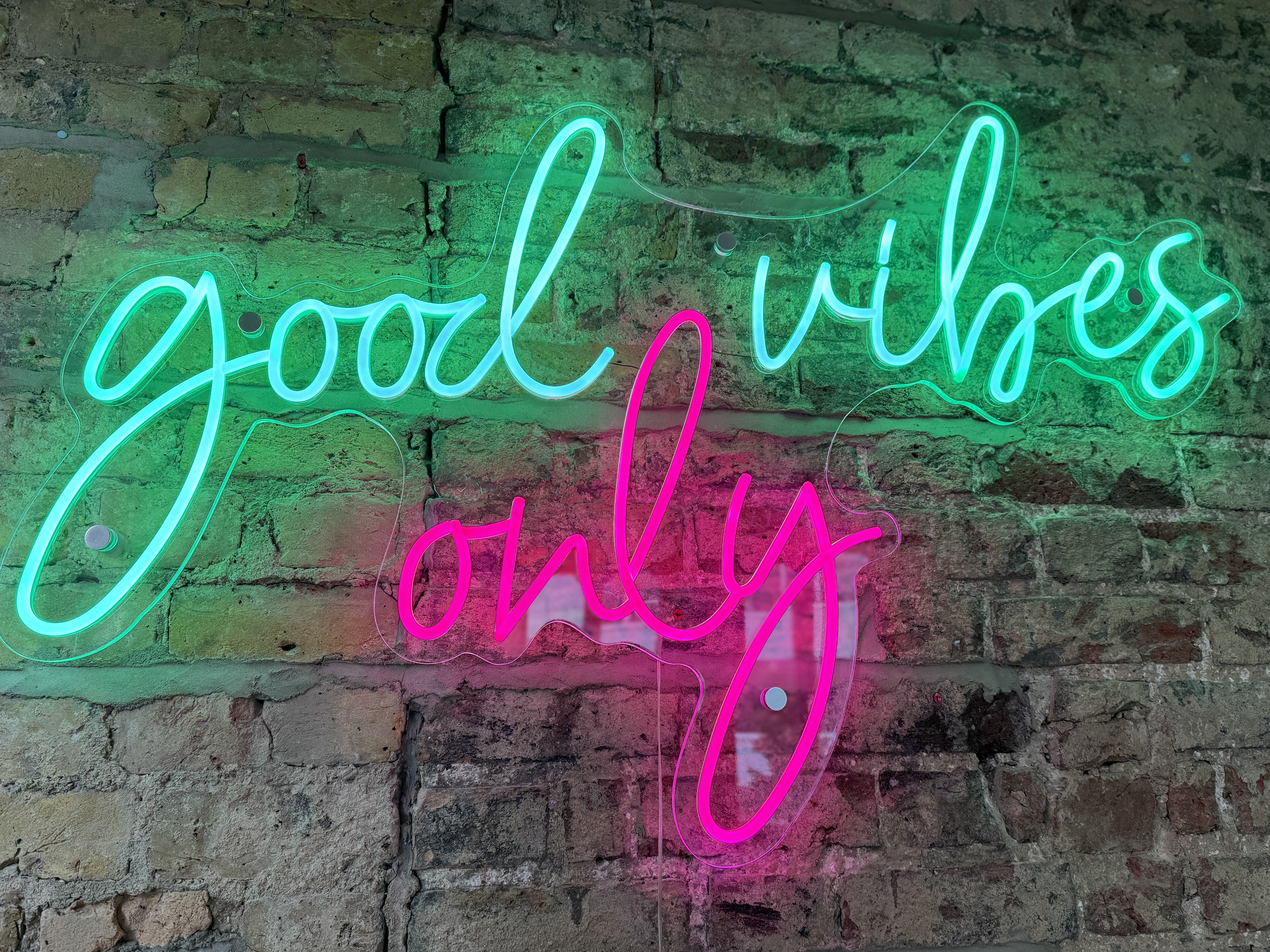 Close-up of a neon wall sign reading ‘good vibes only’ in pink and green lights mounted on an exposed brick wall at The Phoenix pub in London