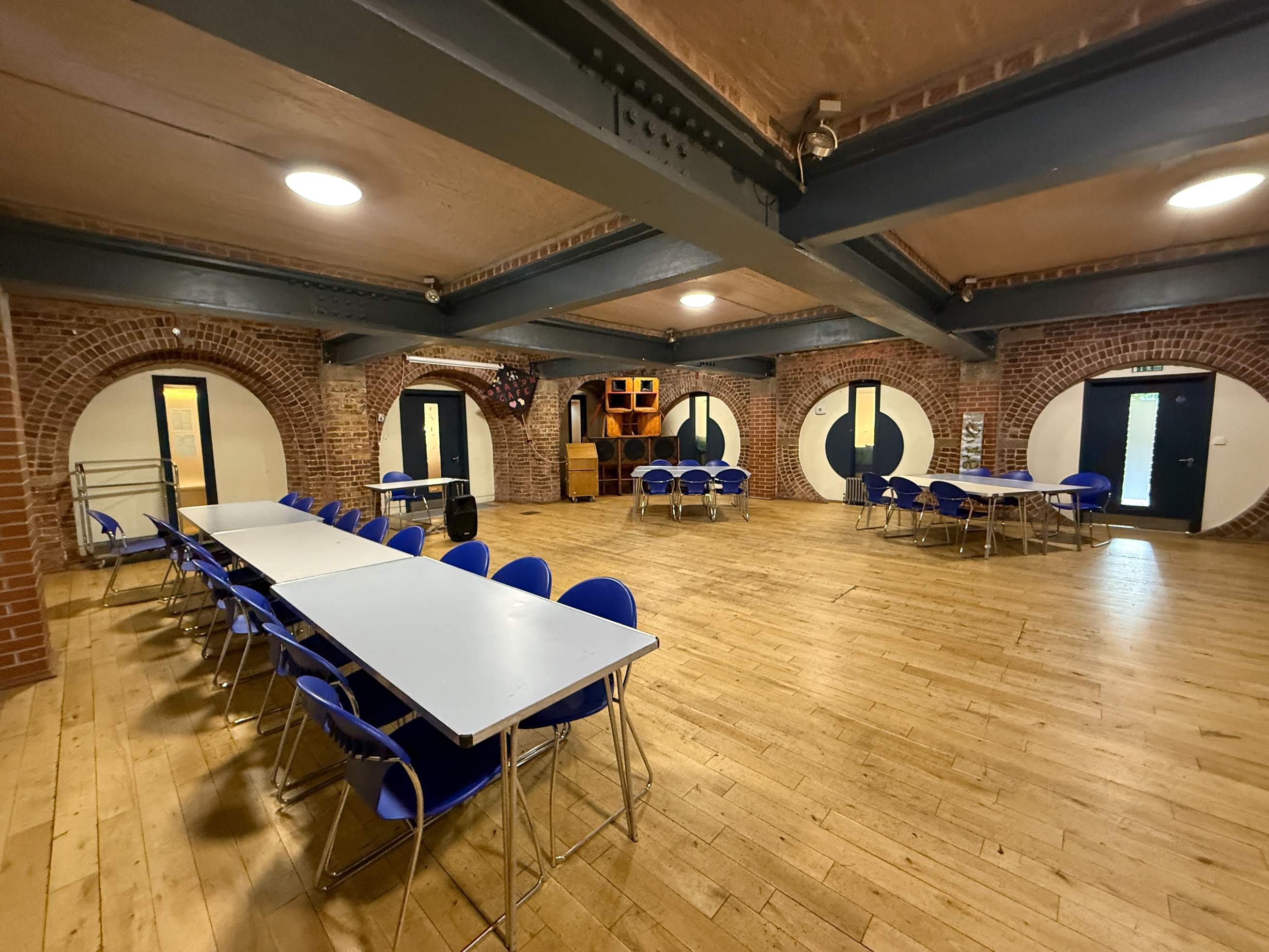 A large, industrial-style community room featuring exposed red brick walls with unique circular arched alcoves. The space has light wood flooring, dark steel ceiling beams, and is furnished with several long white tables surrounded by bright blue chairs.