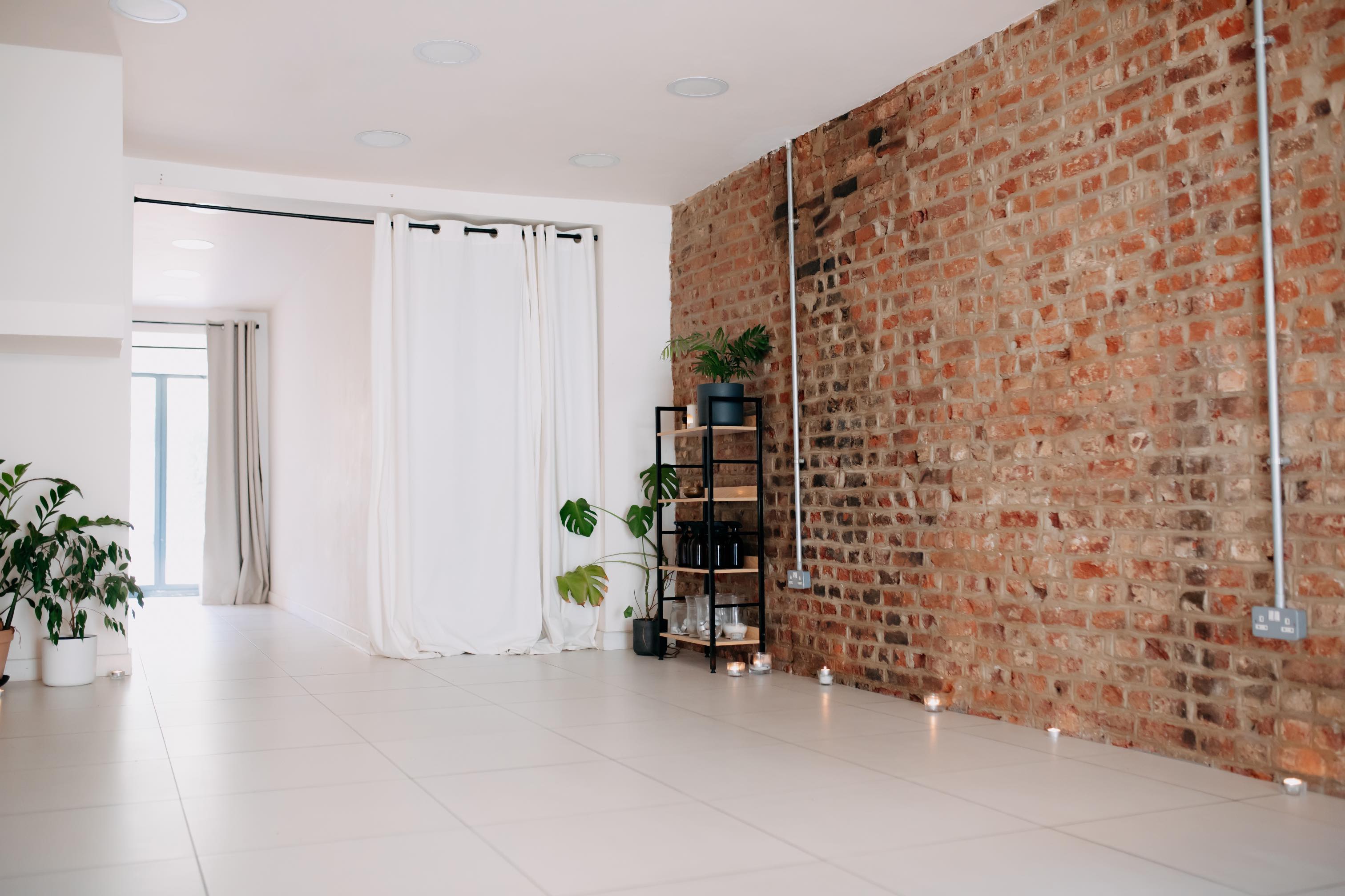 Serene modern studio with white tiled floor, exposed brick wall, potted plants, candles, and decorative shelving unit.