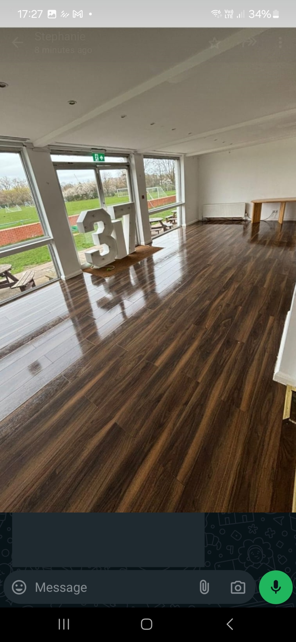  A large, empty function room with a glossy wood floor and floor-to-ceiling windows. The windows look out onto a sports field with a picnic table.