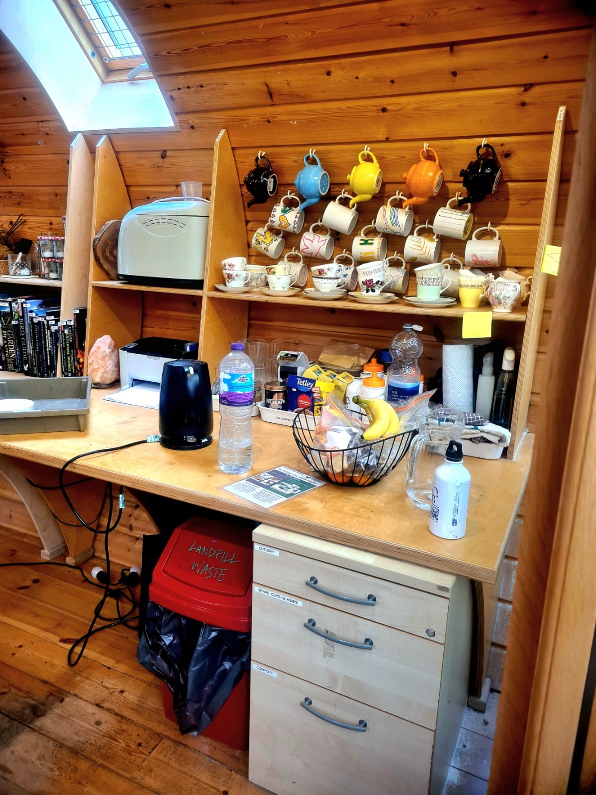 A cluttered wooden countertop with mugs hanging from hooks, a microwave, and a small basket of fruit.