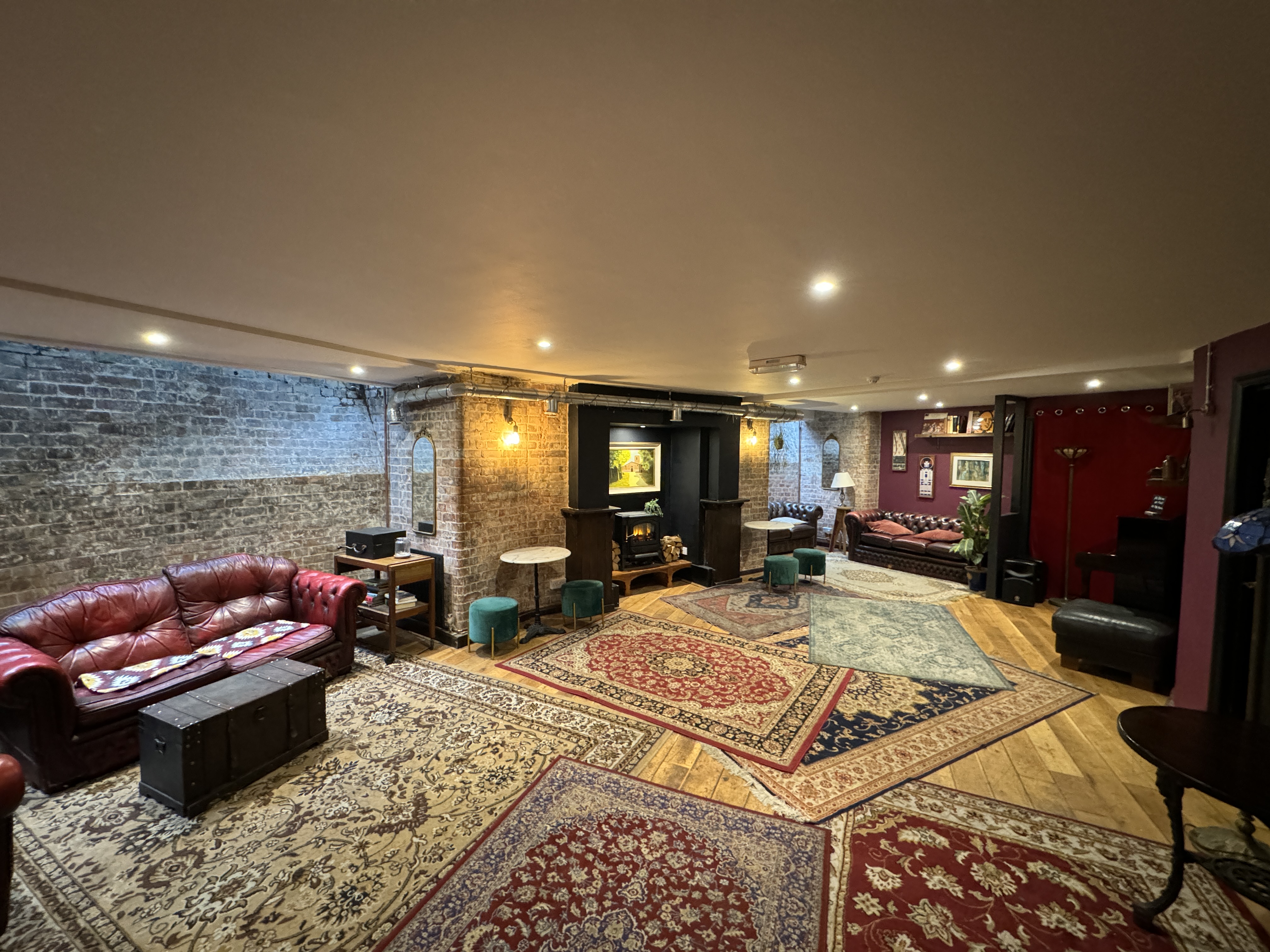 An eclectic basement lounge with exposed brick walls, layered vintage oriental rugs, and warm ambient lighting. The space features tufted red leather Chesterfield sofas, small marble-top bistro tables with teal velvet stools, and a decorative fireplace.