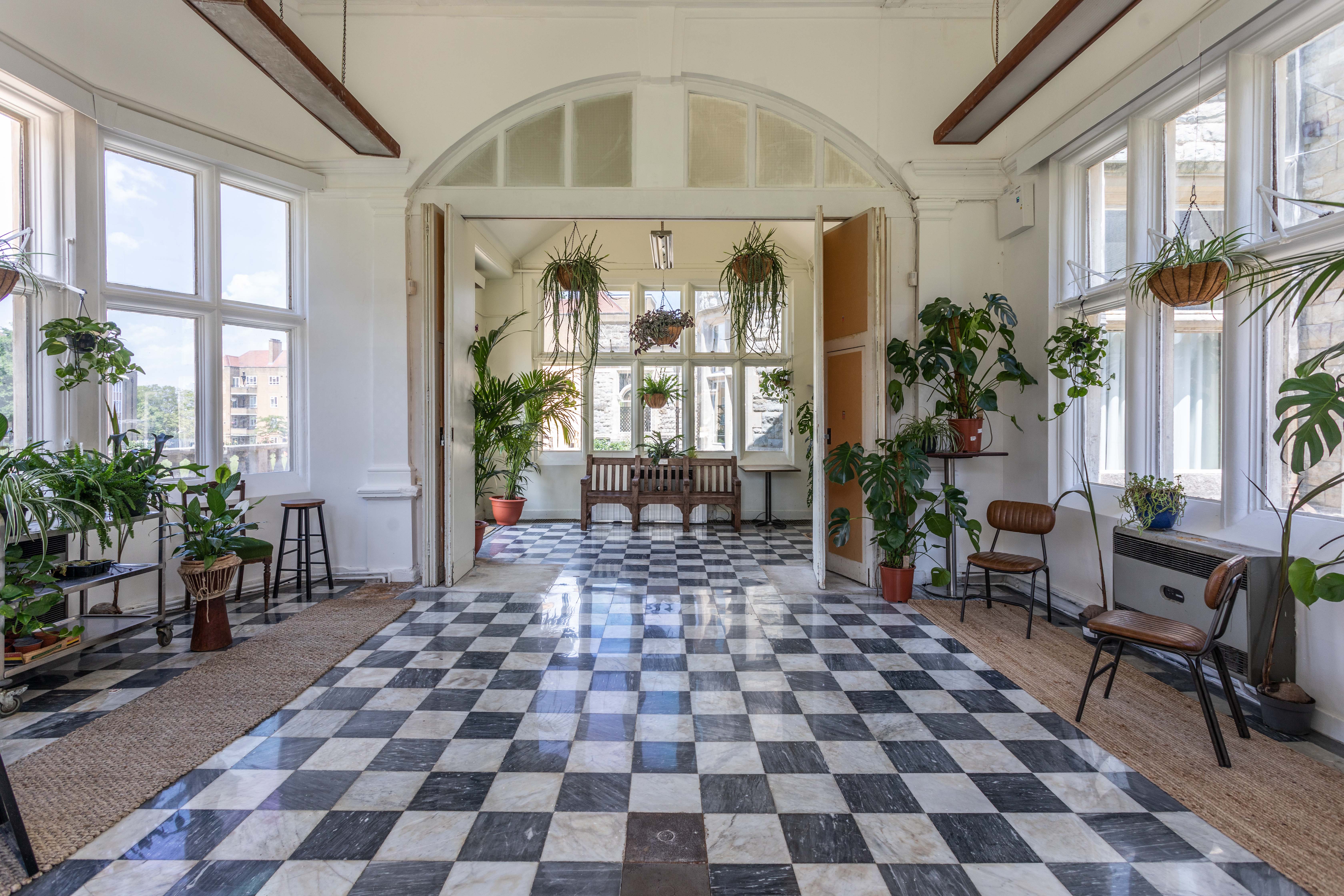 A bright side view of the Palm Court at Kingswood Arts, a 40-capacity space with large windows, a checkered floor, and numerous potted plants.