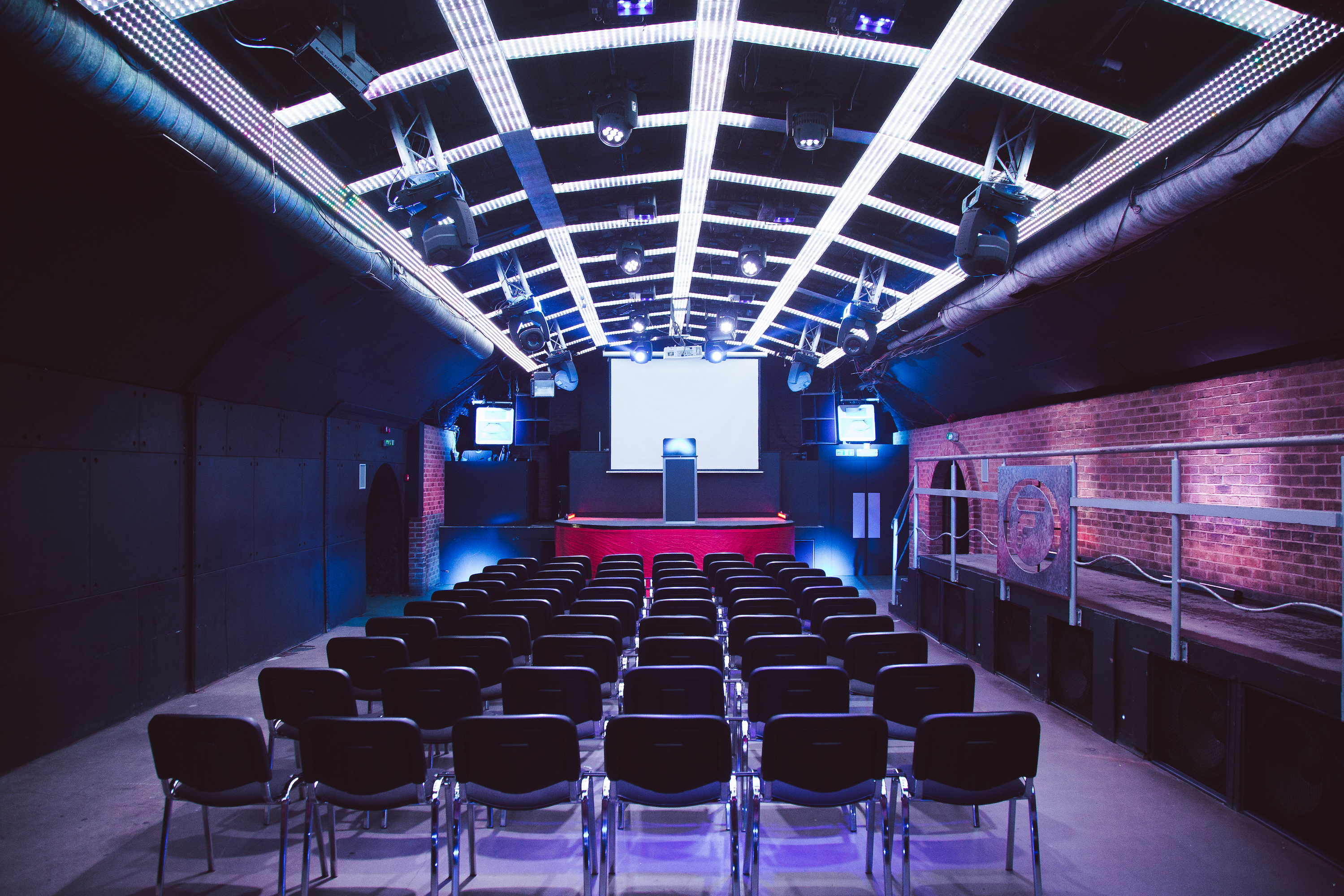 The Fire Main Room at Vauxhall Arches, a 600-capacity venue, is arranged with long picnic benches facing a stage with a gridded light wall.