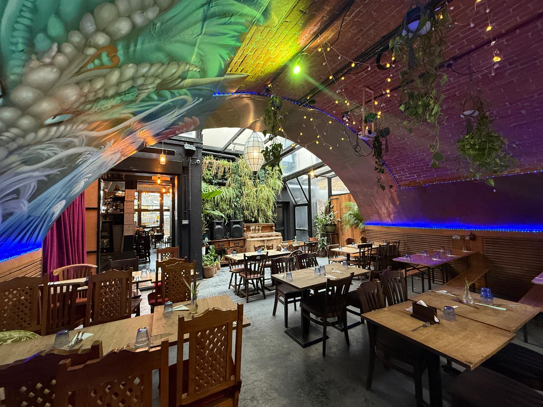 A colorful restaurant with a vaulted ceiling mural, wooden tables, string lights, and a lush living plant wall - available to book via Conveenie.