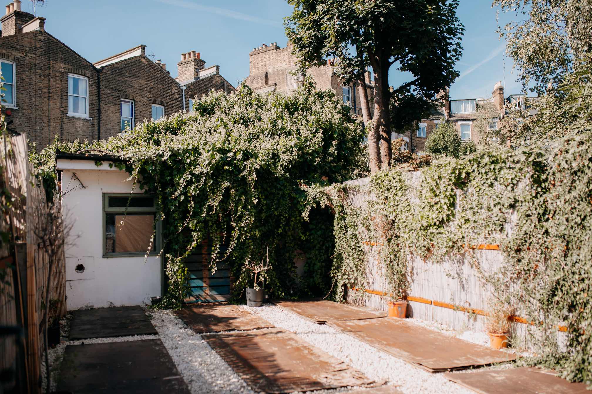 A tranquil outdoor garden space with a small white studio covered in vines, with a mix of wooden decking and white pebbles.