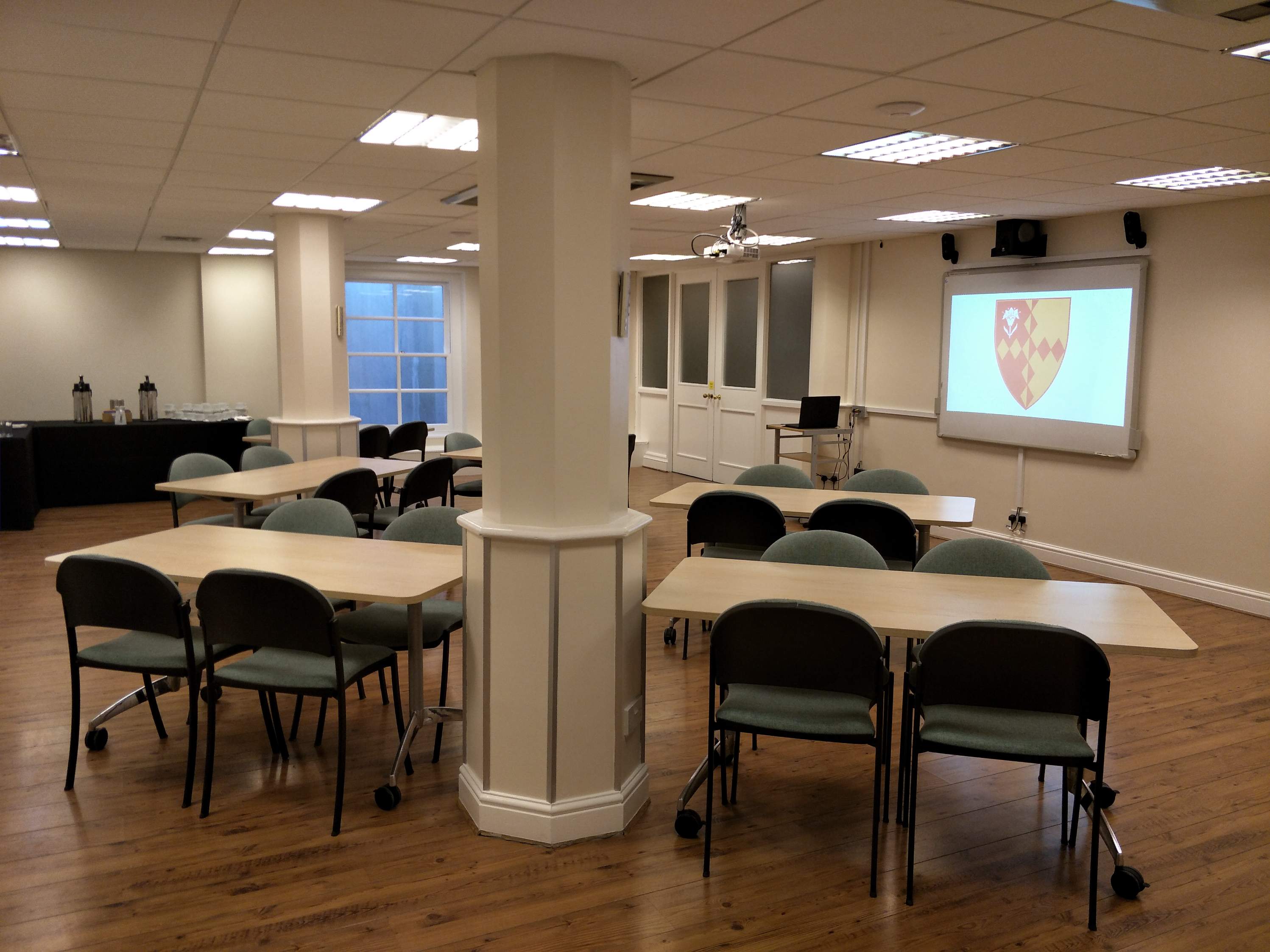 " A classroom-style meeting room with a wooden floor, tables and chairs, a large projection screen, and a large pillar in the center."