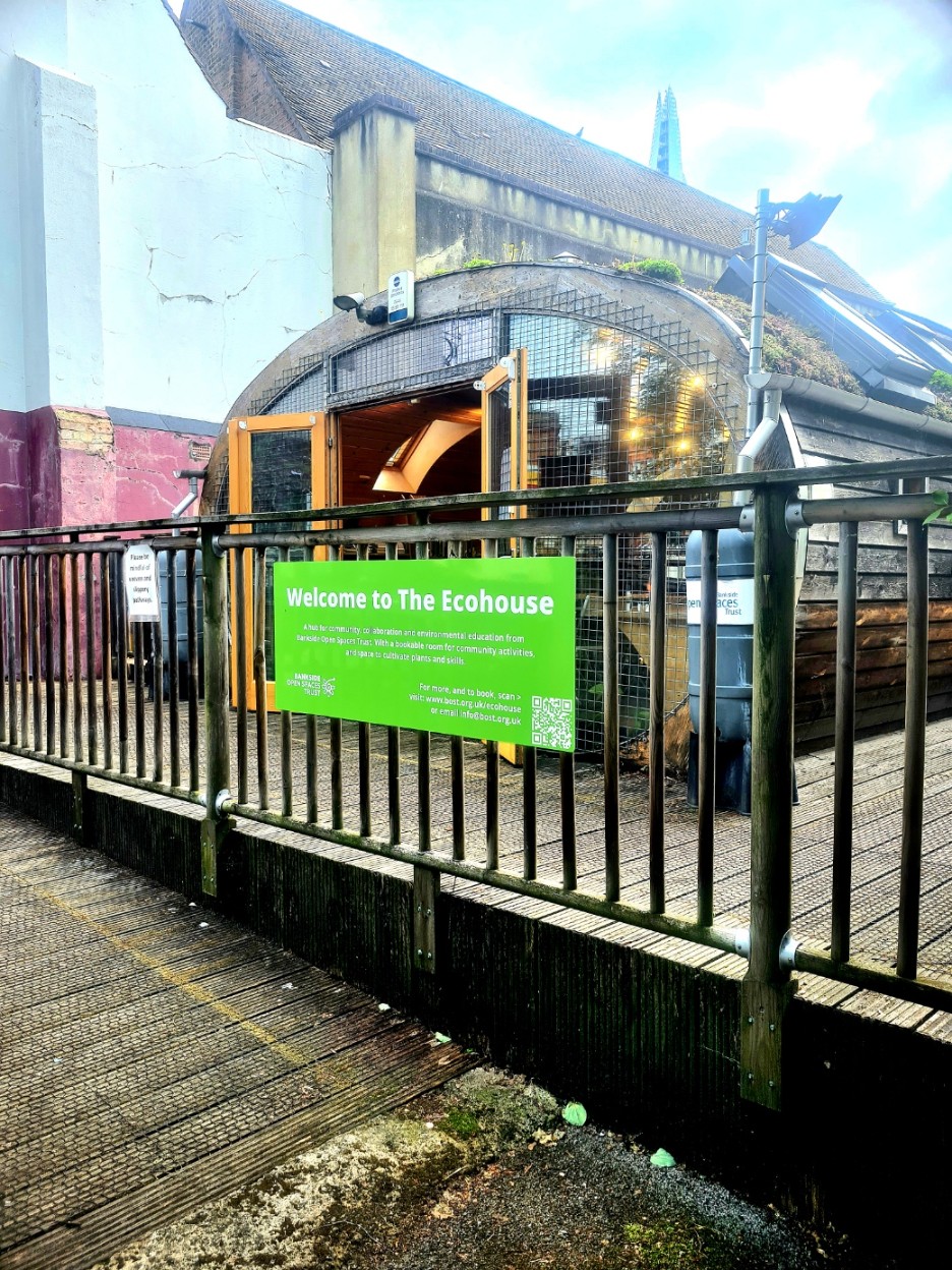 A bright green sign on a metal railing welcomes visitors to "The Ecohouse," a unique, boat-like building with a plant-covered roof.