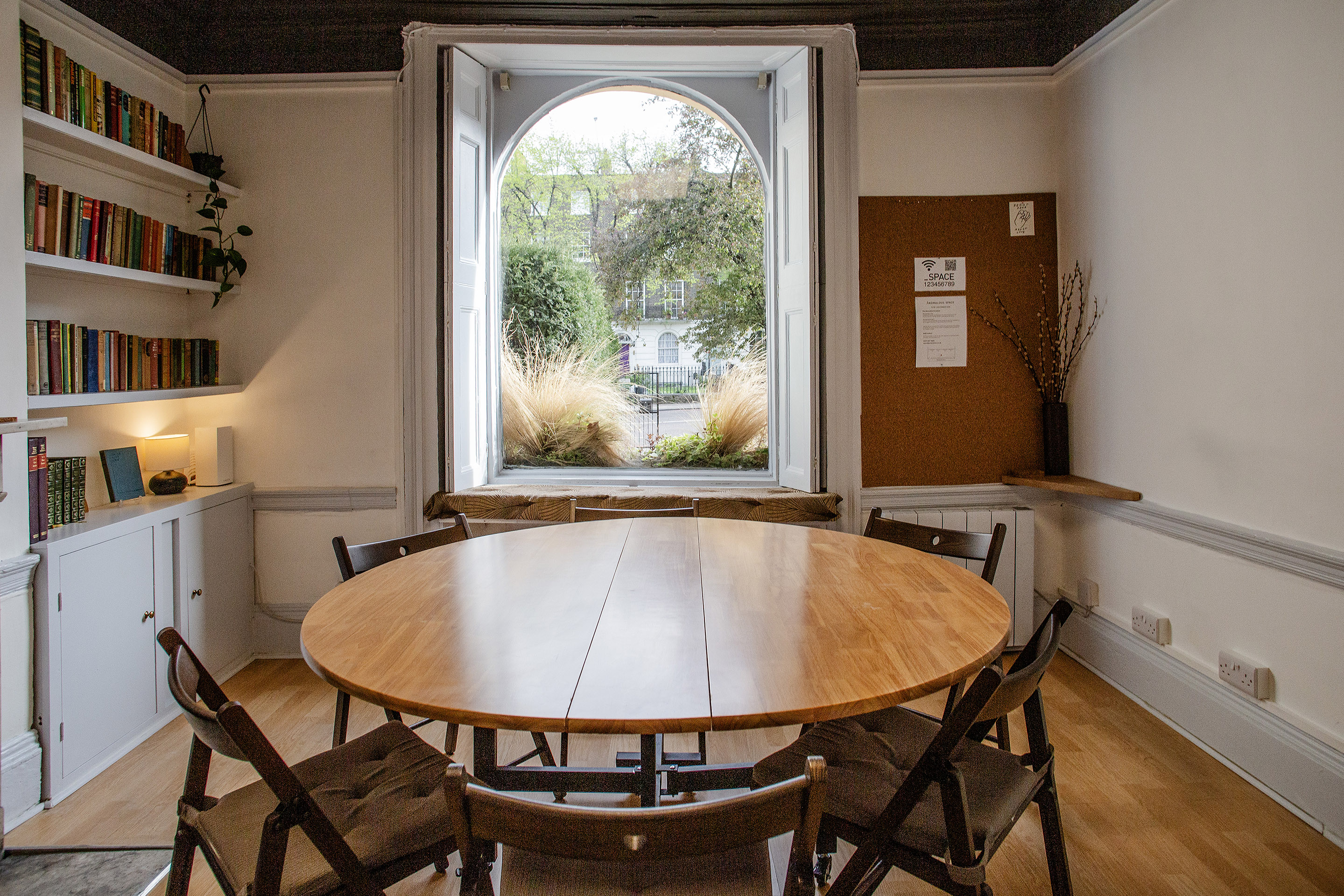 The Study at Anomalous Space, a bright London meeting room for 10, featuring a round wooden table and bookshelf next to an arched window - book via Co