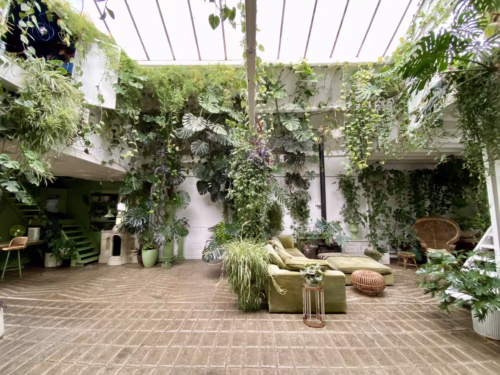"A sun-drenched, spacious indoor sunroom filled with lush tropical plants covering the walls and hanging from a glass ceiling, featuring a green velvet sofa and patterned stone flooring."