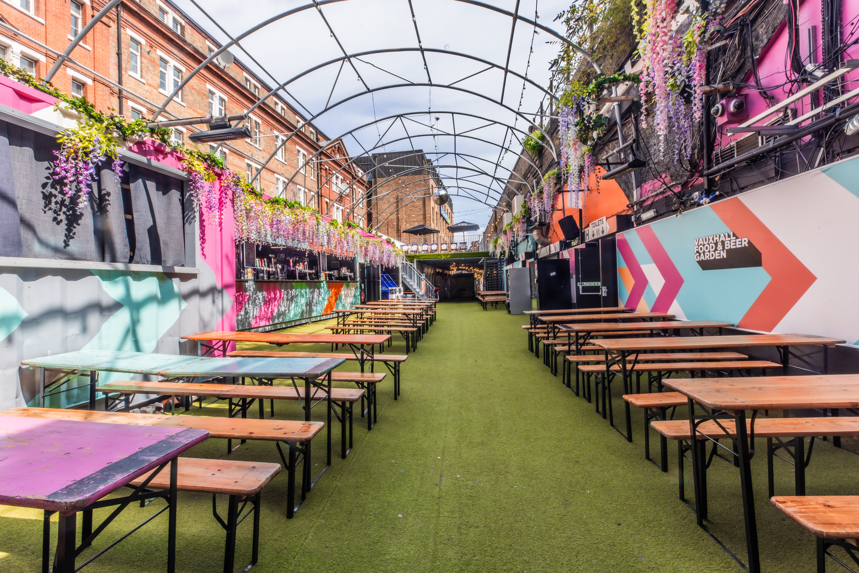 The spacious Vauxhall Food and Beer Garden for 800, bookable through Conveenie, features picnic tables, an arched roof, and colorful vendor stalls.