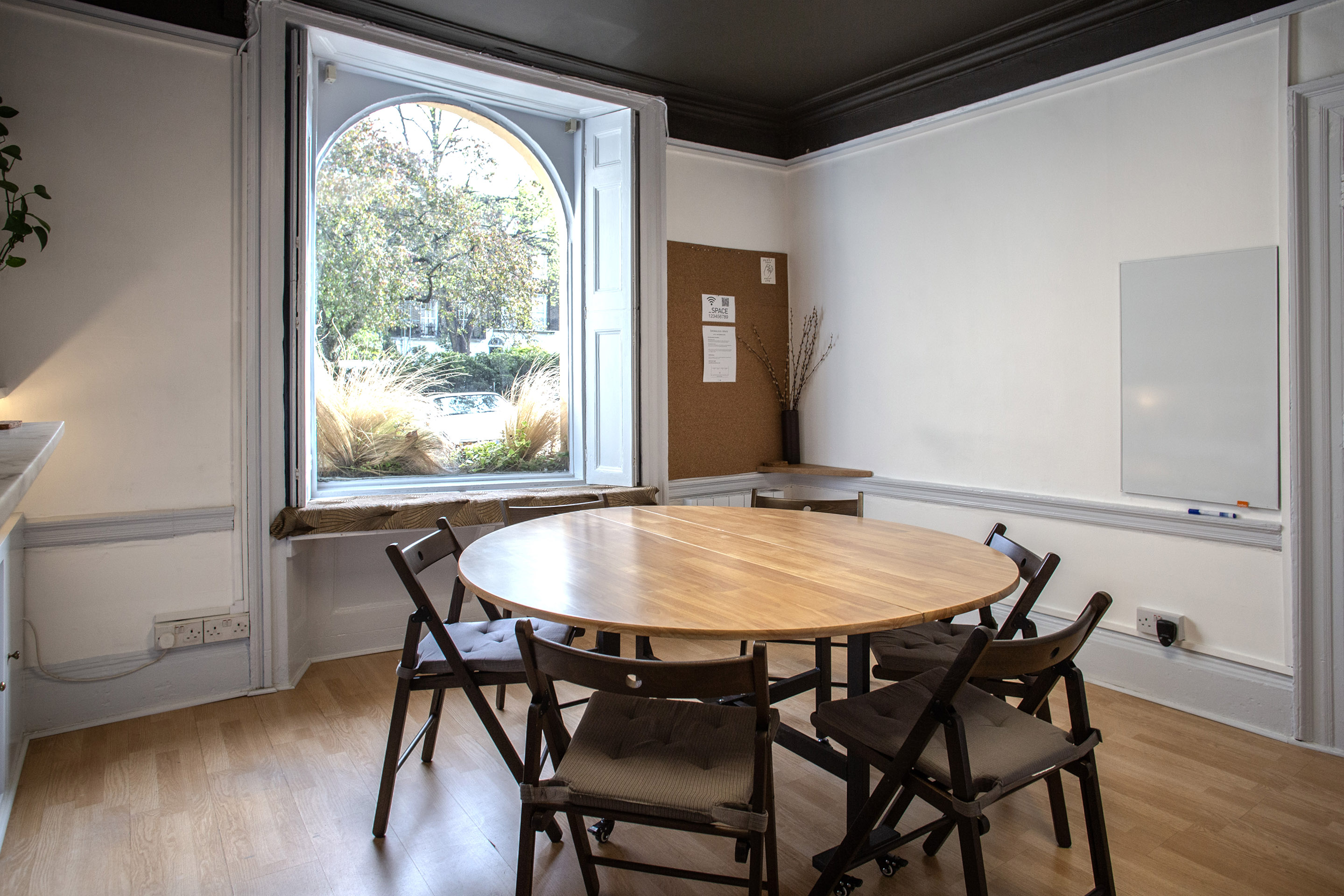 The Study at Anomalous Space, a bright London meeting room hire for 10, featuring a round wooden table, white bookshelf, and arched window view.