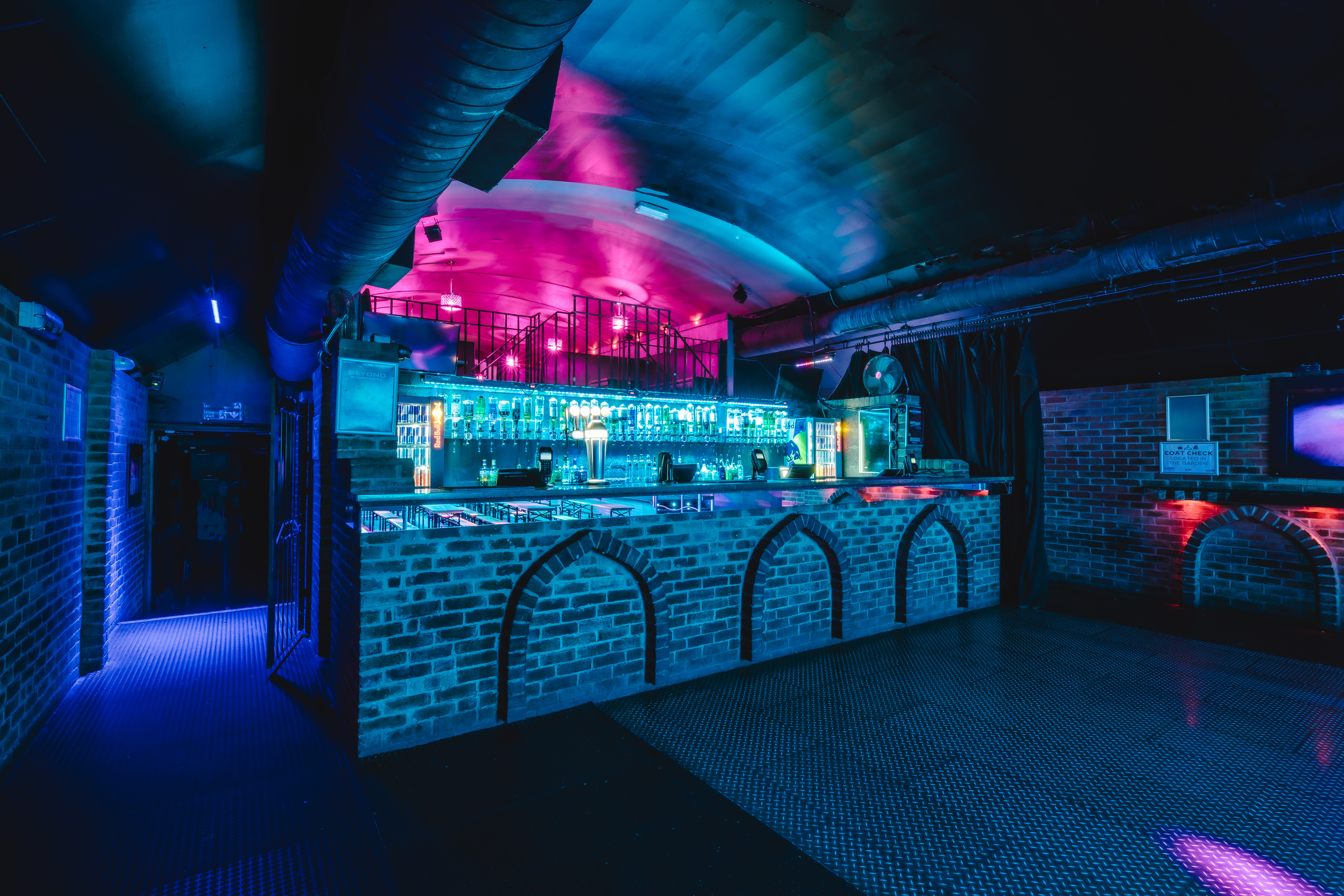 A stylish bar at Vauxhall Arches, a 600-capacity venue, features an exposed brick front with arches, illuminated by cool blue and pink lights.