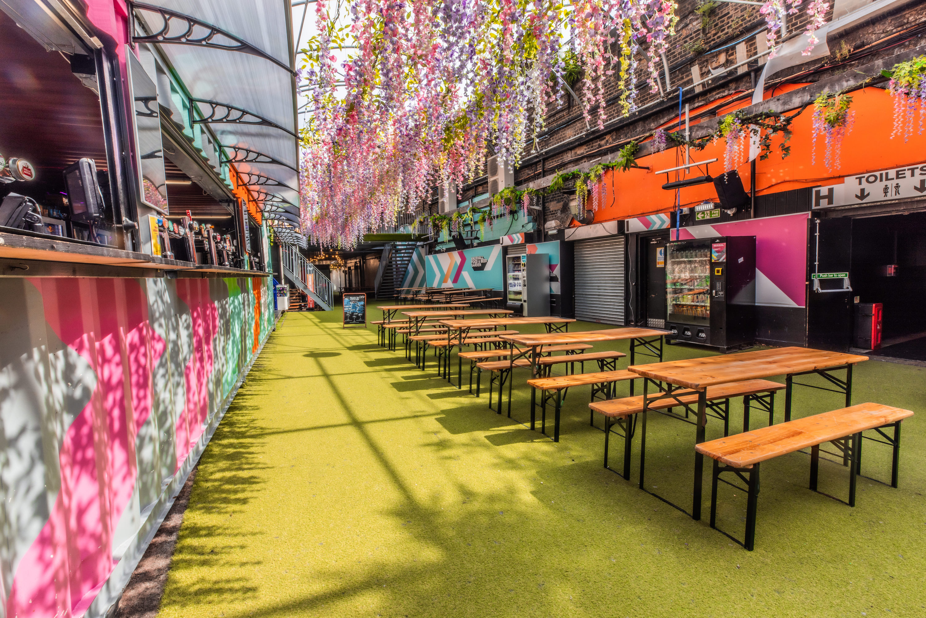 The Vauxhall Food and Beer Garden, for 800 and listed on Conveenie, features green turf, picnic tables, and a container bar under hanging wisteria.
