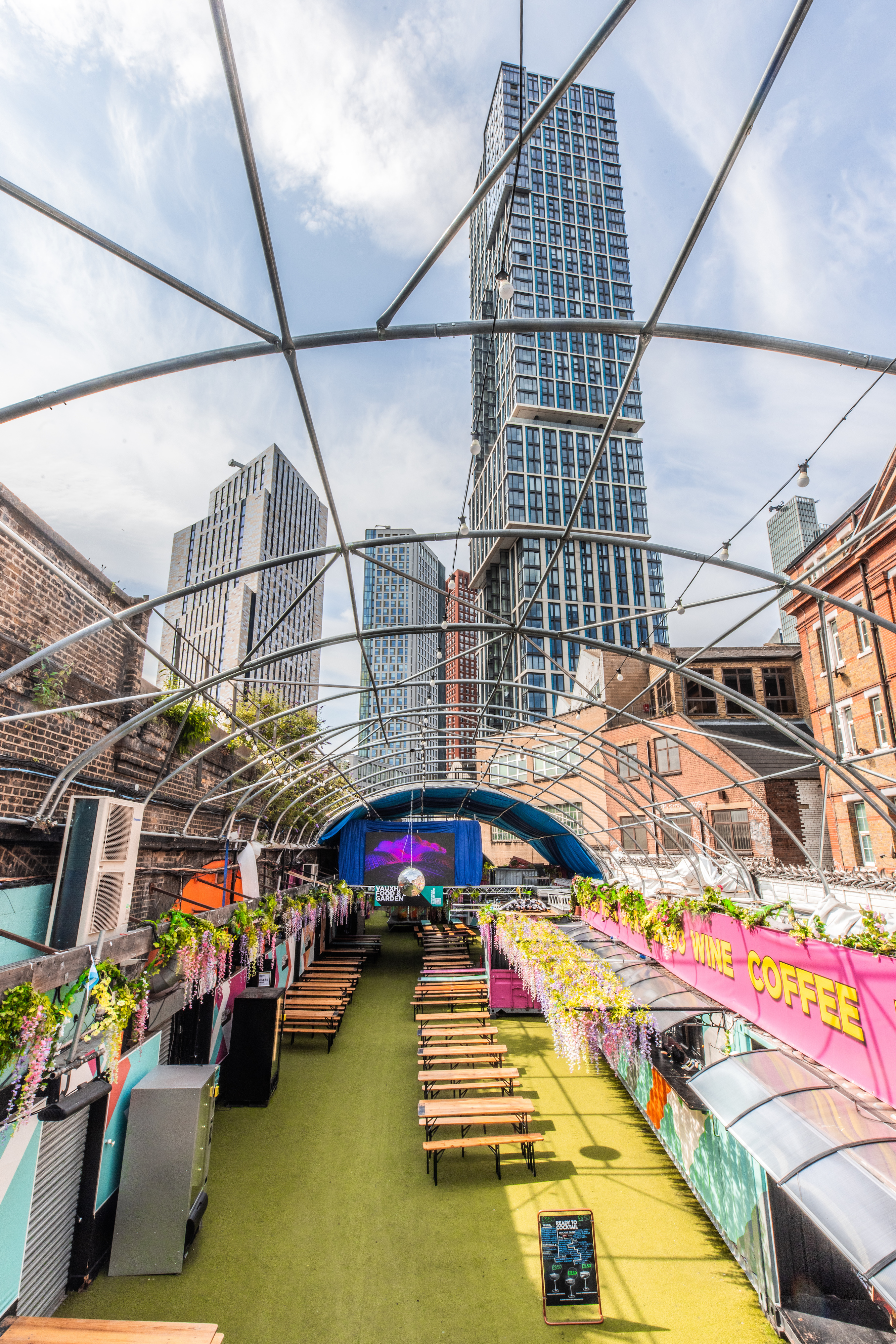 The main bar at Vauxhall Food and Beer Garden, a venue for 800, features numerous beer taps, colorful decor, and is adorned with hanging flowers.