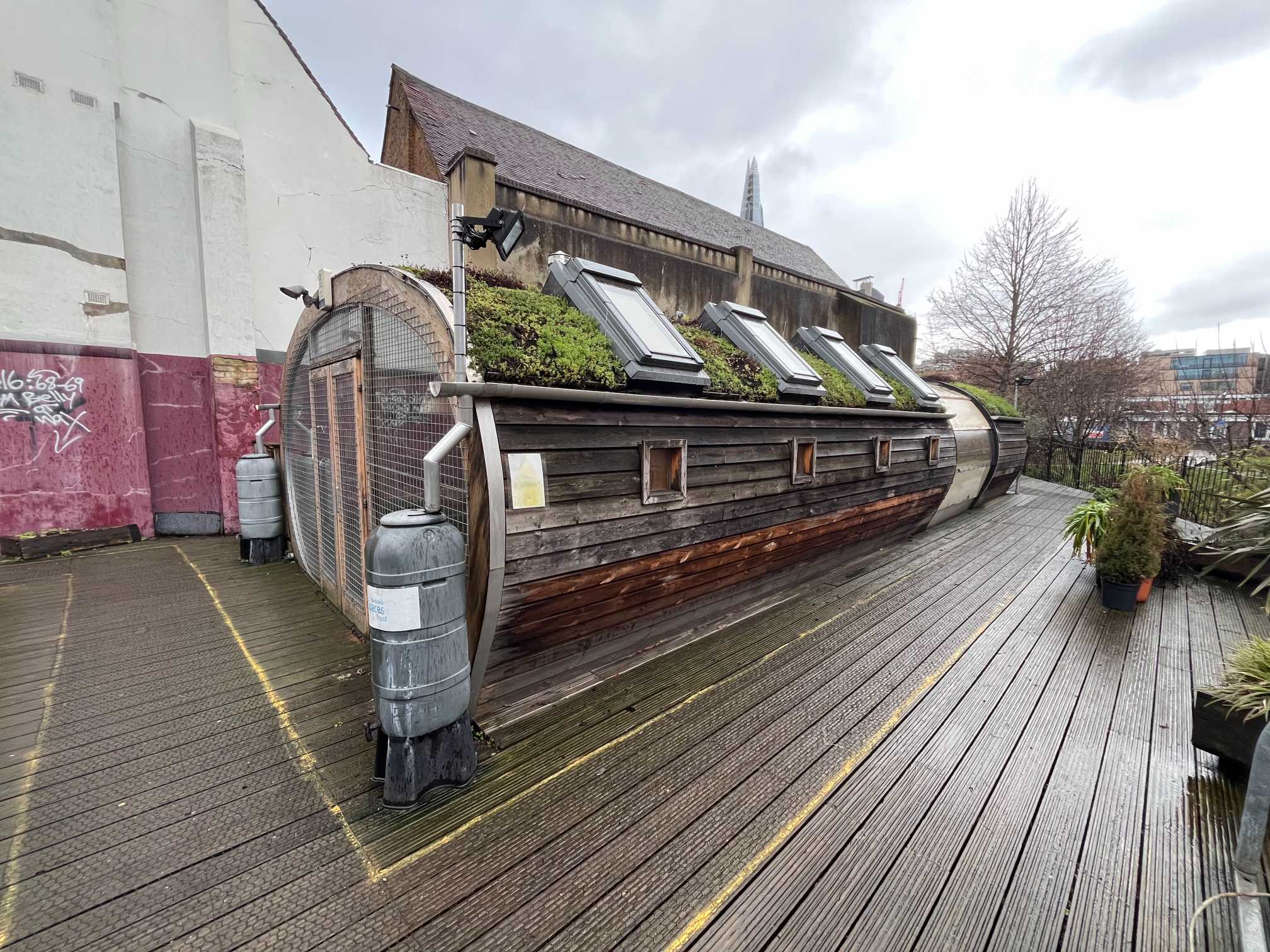 A narrow, wooden building with a rounded exterior, a green roof, and multiple skylights, all built on a wooden deck - available to book via Conveenie.
