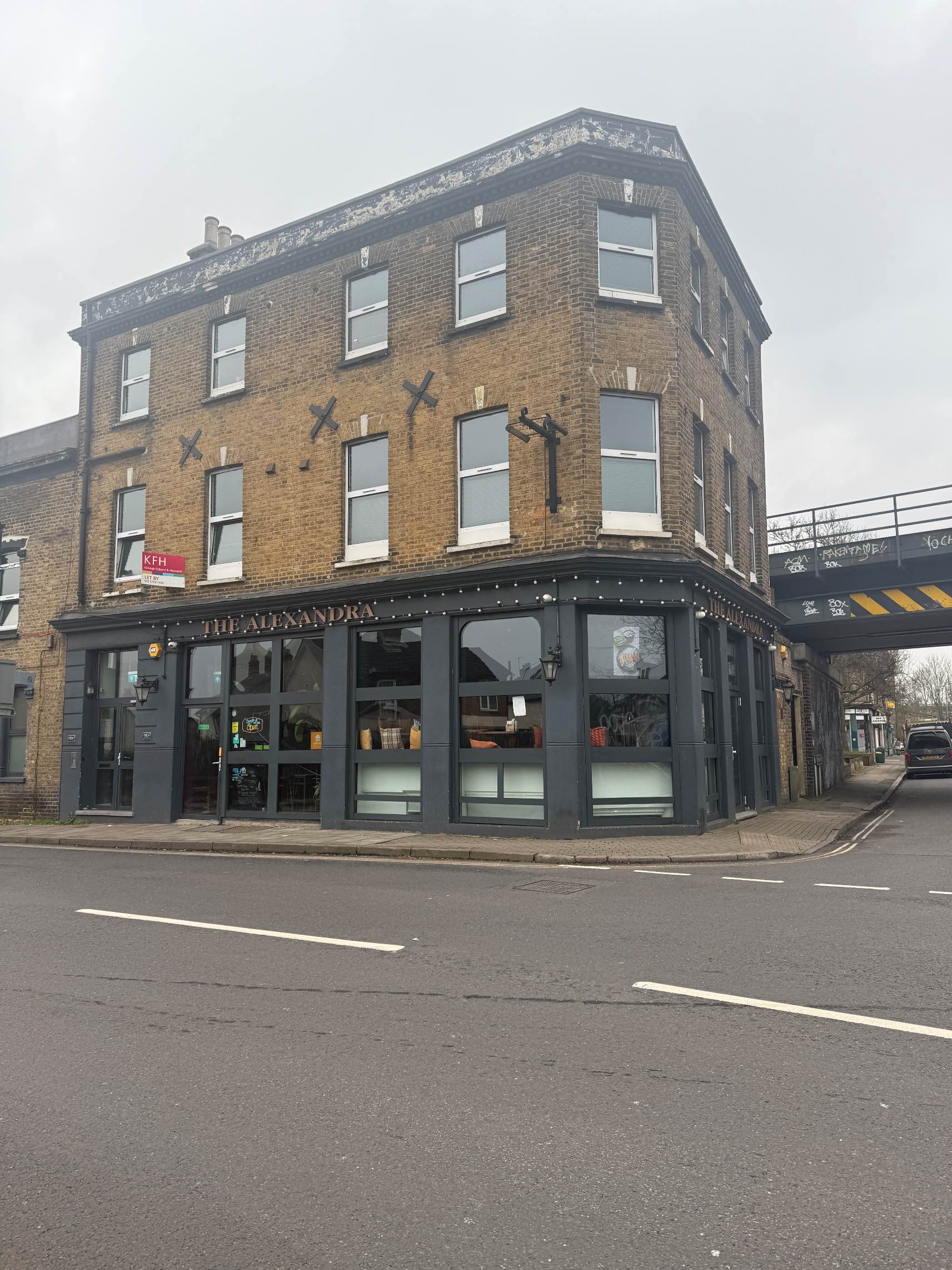A large, three-story brick corner building with the pub's name, "The Alexandra," visible above a dark grey facade with large windows.