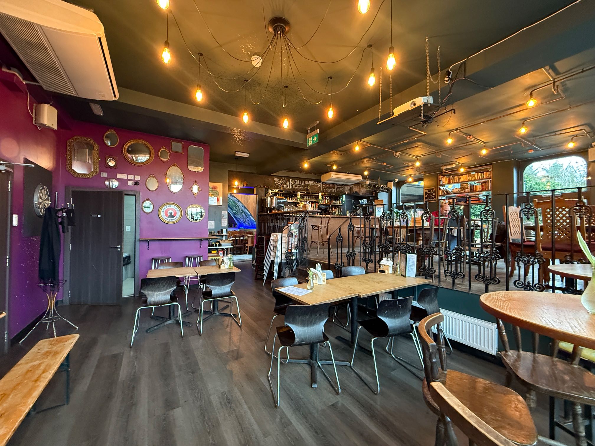 A lively pub interior with various wooden tables and chairs, a mix of mirrors on a purple accent wall, and a bar area with antique-style furniture.