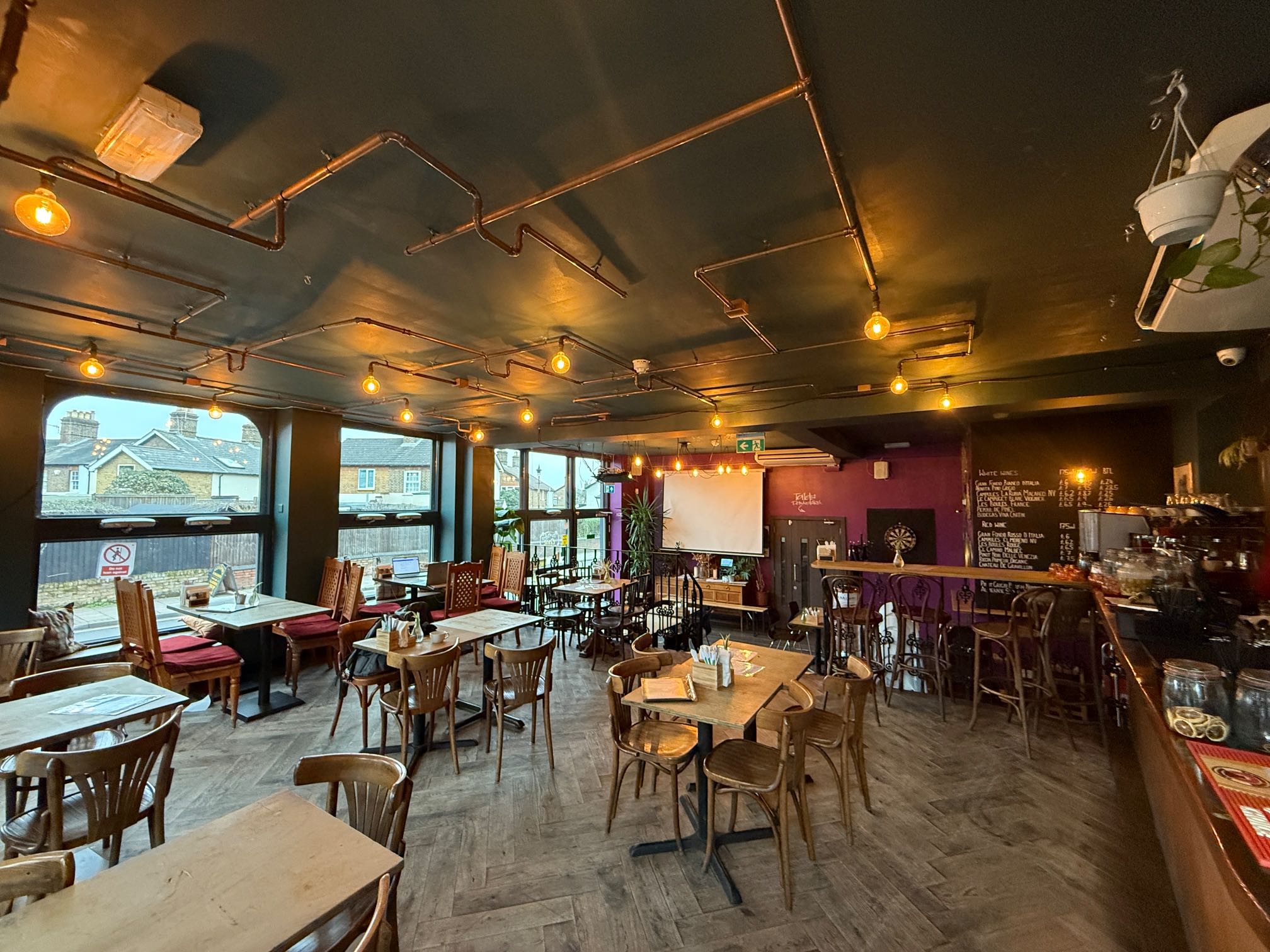 Pub interior with a dark green ceiling, exposed copper pipes, and a mix of rustic wooden tables and chairs, featuring large windows and a bar area.