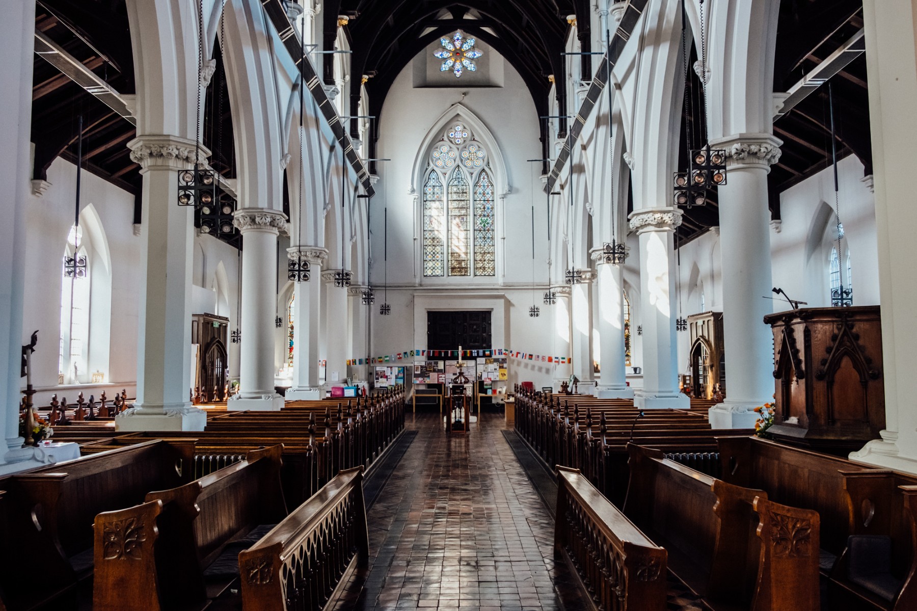 The interior of a traditional church featuring long wooden pews flanking a central aisle that leads toward an ornate stained-glass window. High vaulted ceilings with dark wood beams are supported by massive white stone pillars, and natural light streams through gothic-style windows.