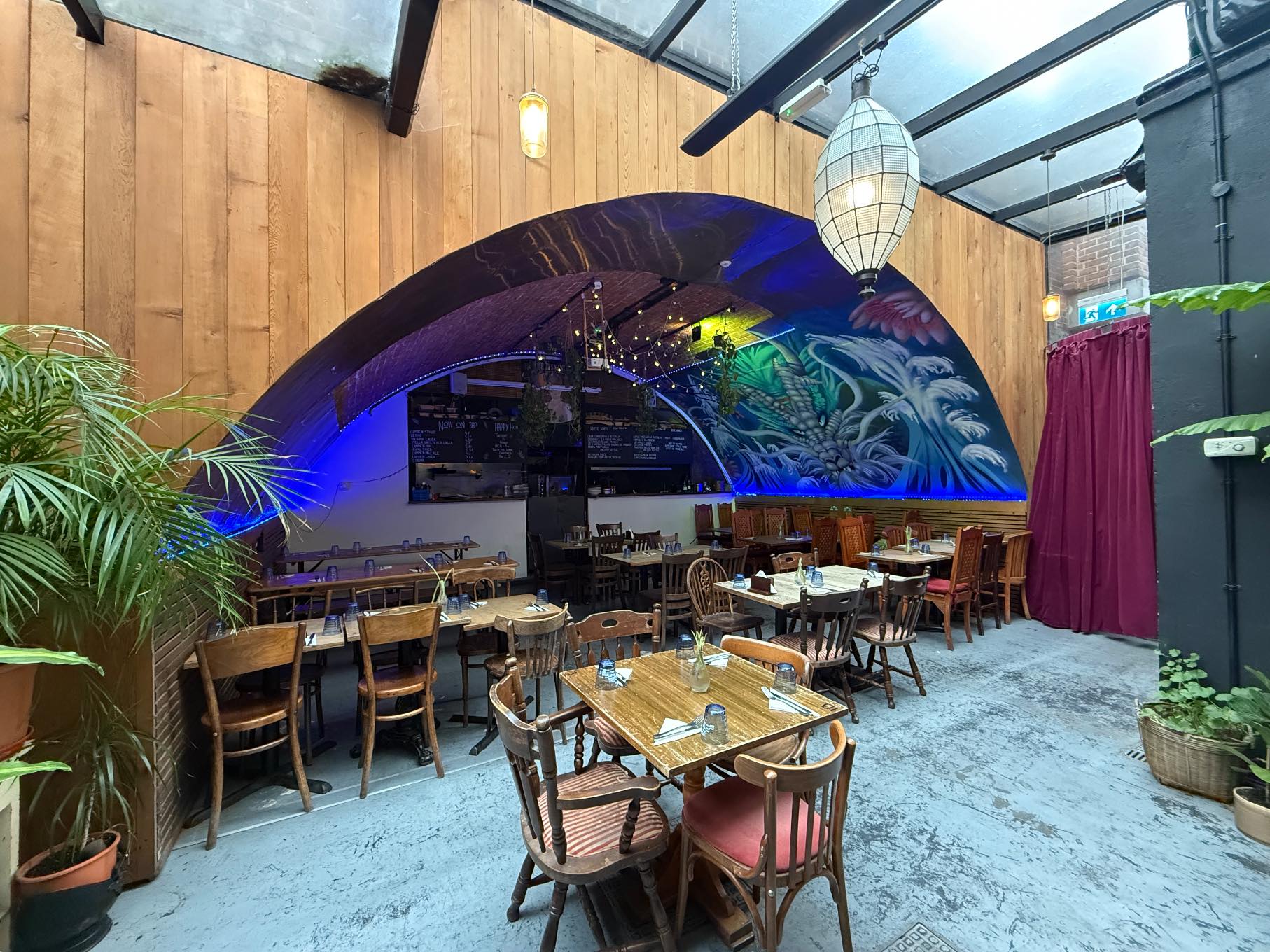 A restaurant with a vibrant mural on a vaulted ceiling, a mix of wooden tables and chairs, and a glass ceiling with a large lantern.