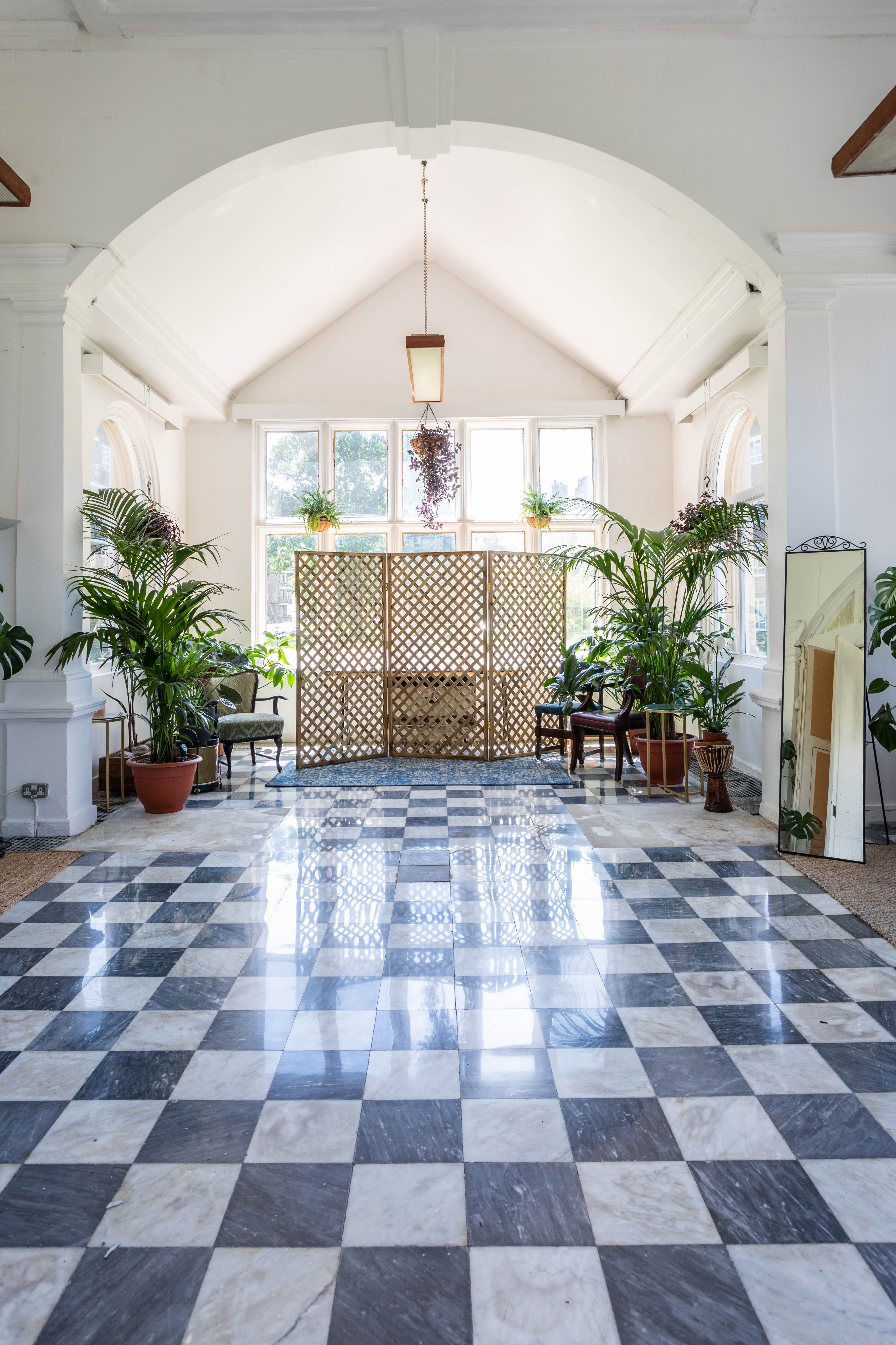 The elegant Palm Court at Kingswood Arts, listed on Conveenie, features a black and white checkered floor, large windows, and lush plants for 40.