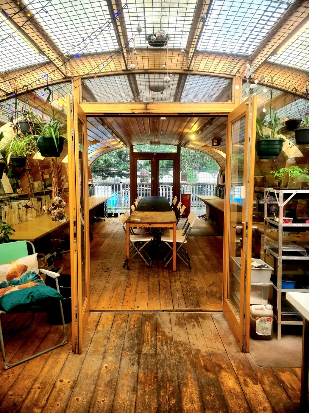 A long, wooden meeting room with a vaulted ceiling, a glass roof, a large table with folding chairs, and hanging plants.