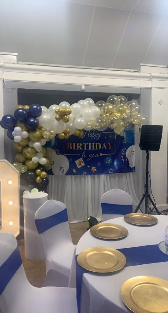Function room with “Happy Birthday” banner, blue-white-gold balloon arch, and tables set with gold placemats and blue runners.