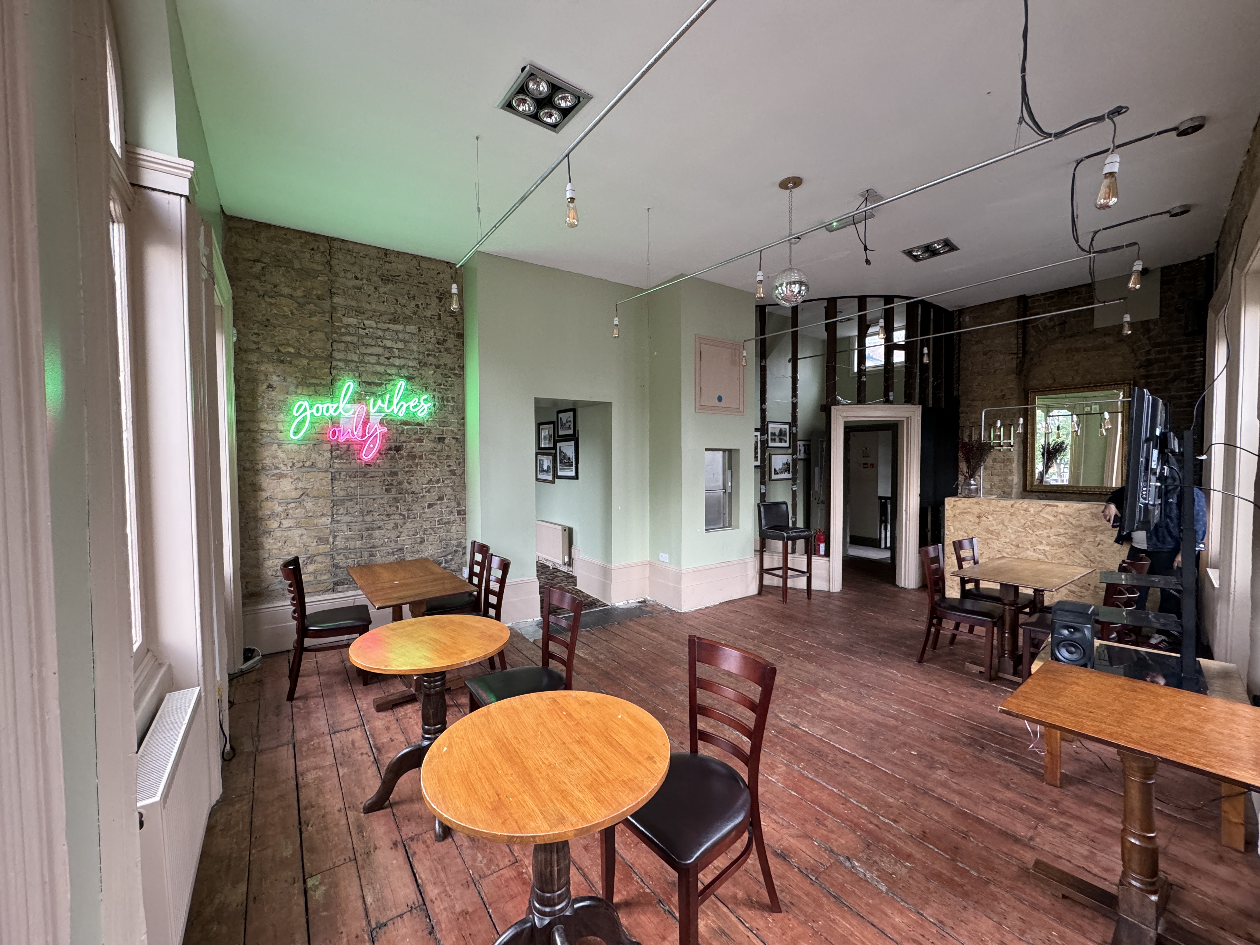 View across the function room: tables and chairs, exposed brick wall, neon wall sign reading ‘good vibes only’, and large windows.